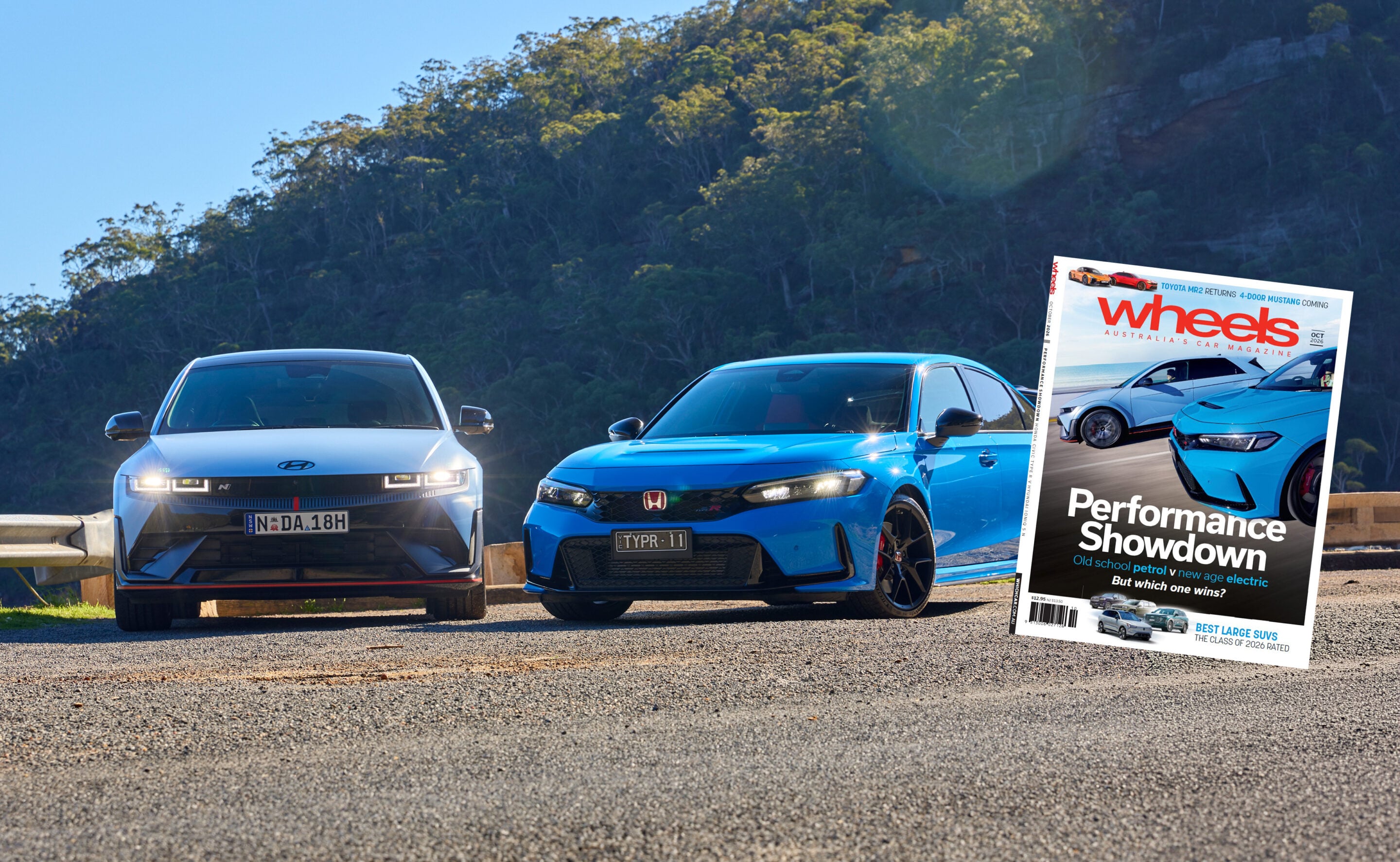 Two modern cars parked on a gravel surface with a forested hillside behind; a tilted Wheels magazine cover overlays the right side.