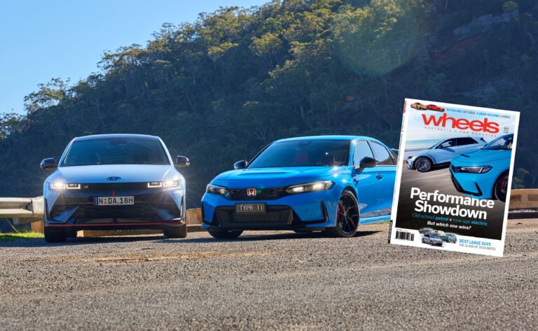 Two modern cars parked on a gravel surface with a forested hillside behind; a tilted Wheels magazine cover overlays the right side.