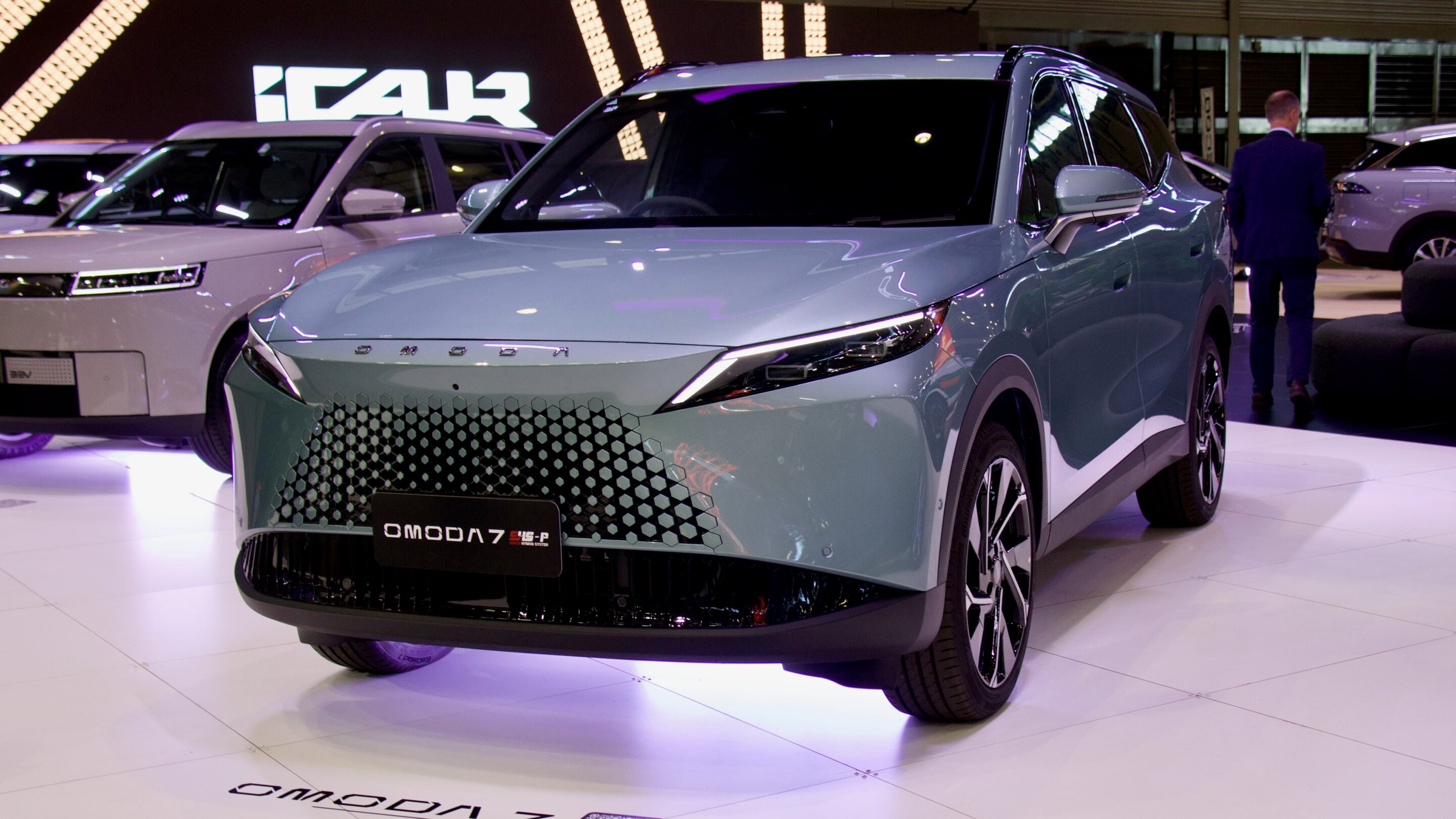 Front view of a light blue OMODA 7 SUV on a showroom floor, with a white SUV and a man in a suit in the background.