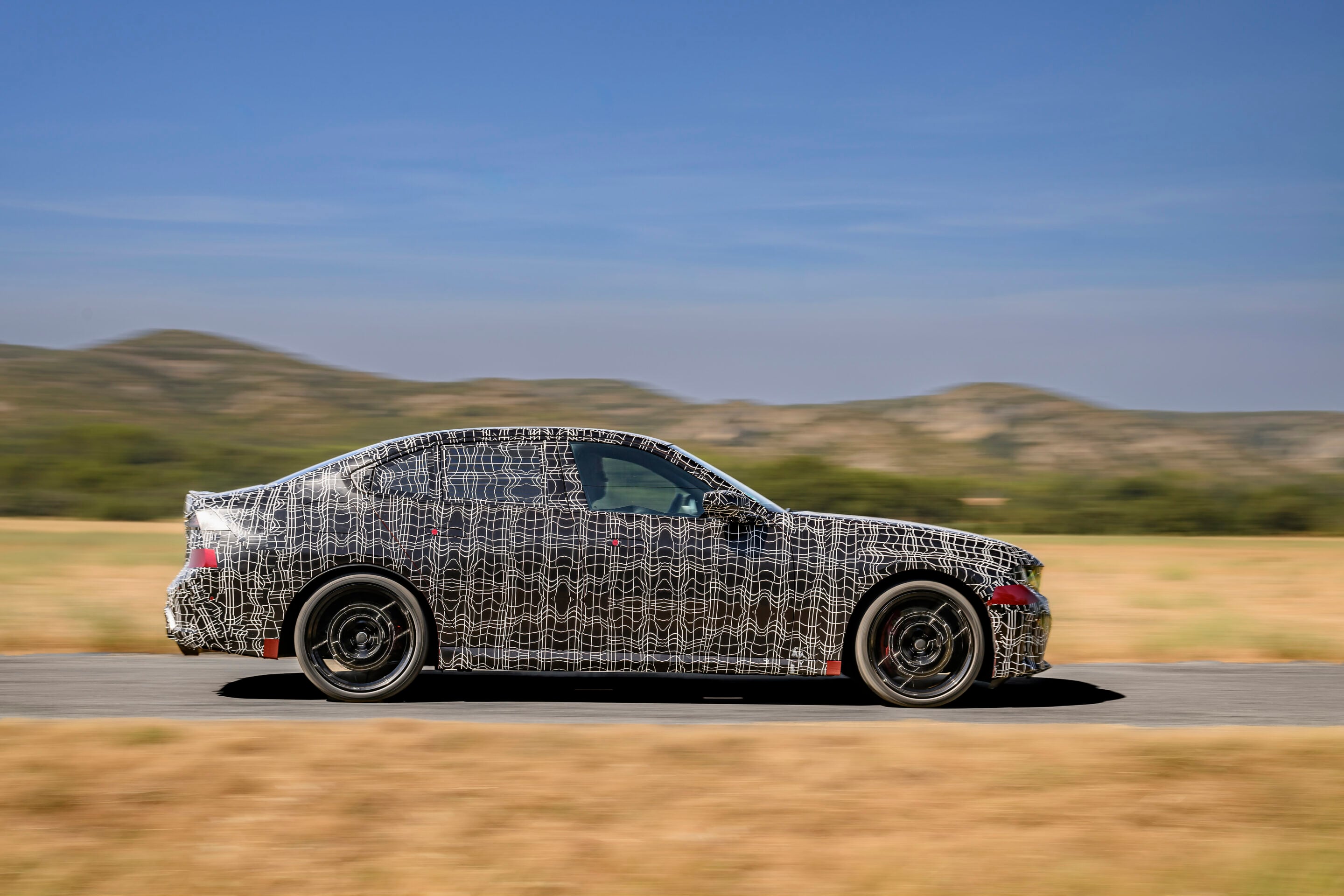 Camouflaged prototype car with white lattice wrap driving on a highway, desert hills in the background