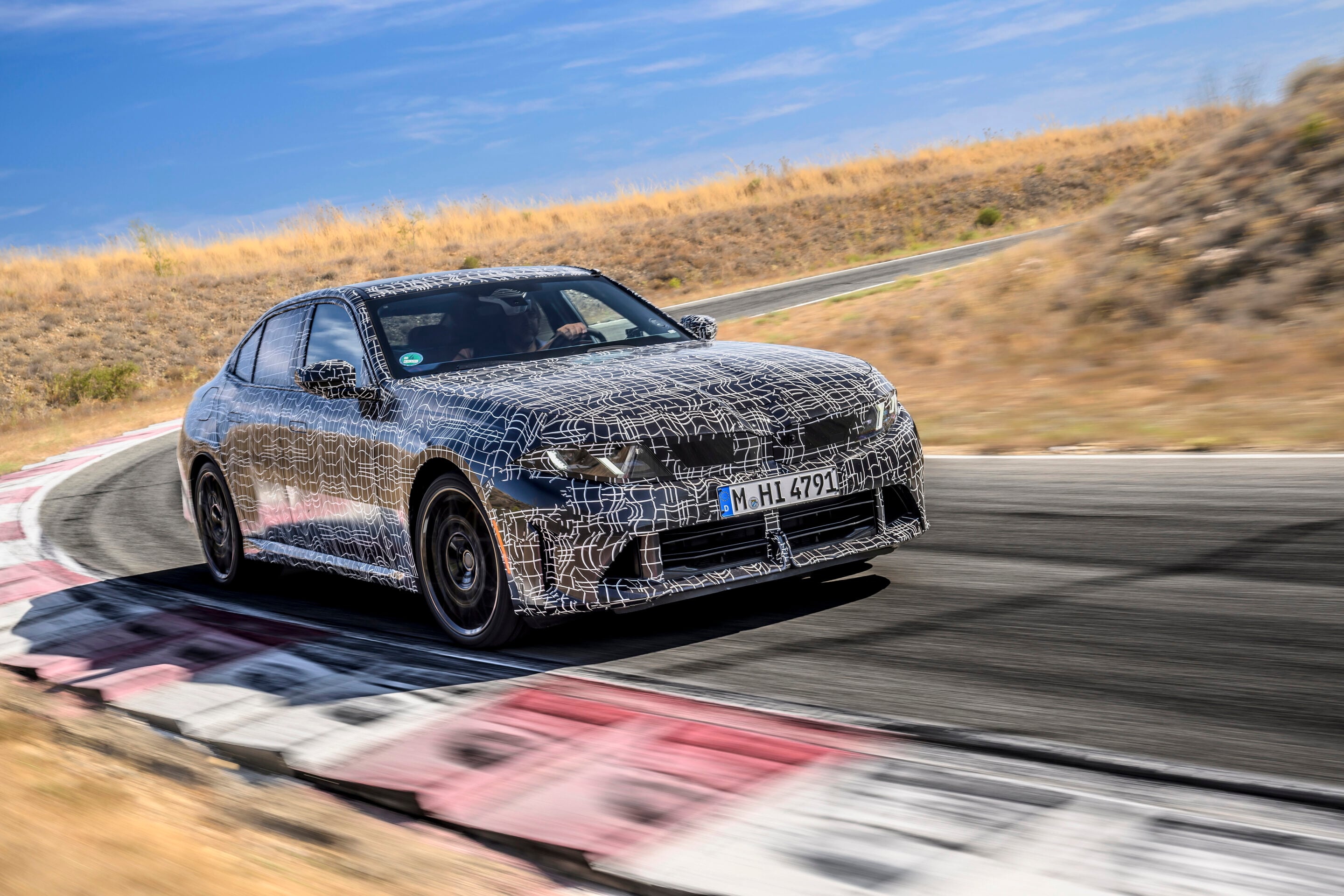 Camouflaged prototype car at speed on a race track, blurred background and curving road with red-and-white barriers, sunny desert surroundings.