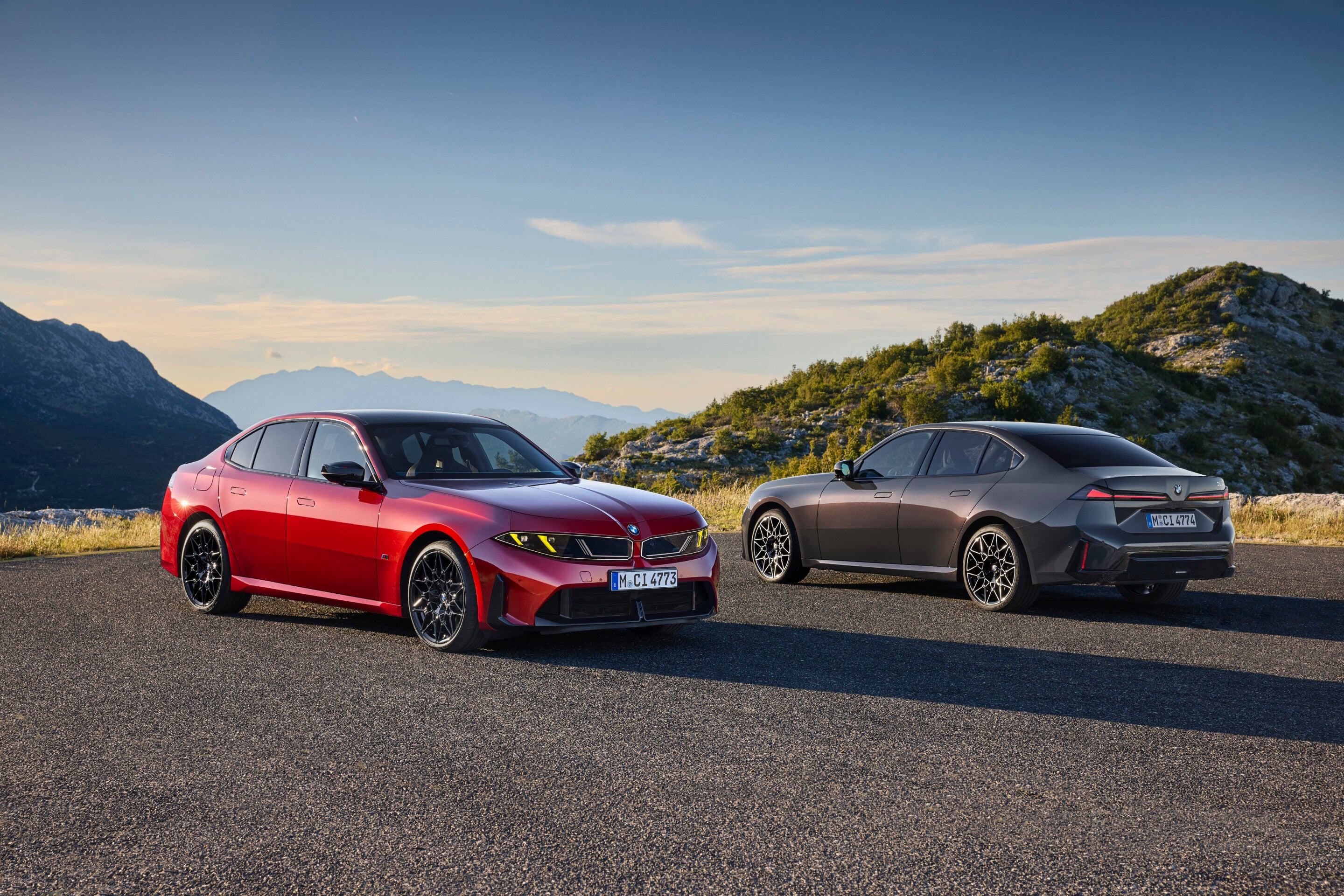 Two BMW cars parked on a mountain road: a red sedan on the left and a gray coupe on the right, with hills and blue sky behind.