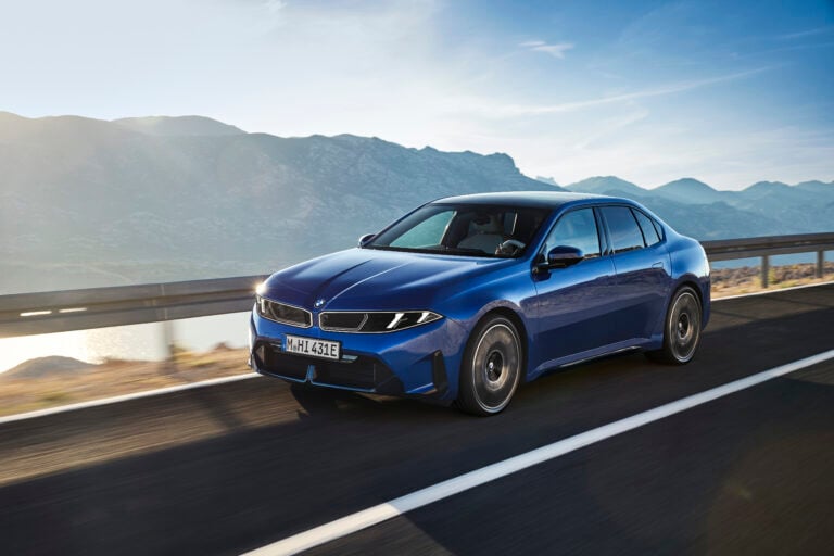 Blue BMW sedan in motion on a mountain highway, guardrail to the side and alpine peaks in the distance.