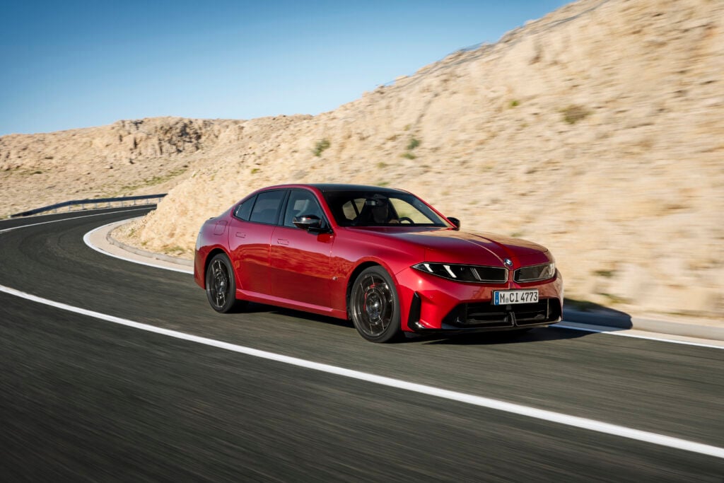 Red BMW sedan driving on a winding mountain road beside rocky cliffs under a clear blue sky.