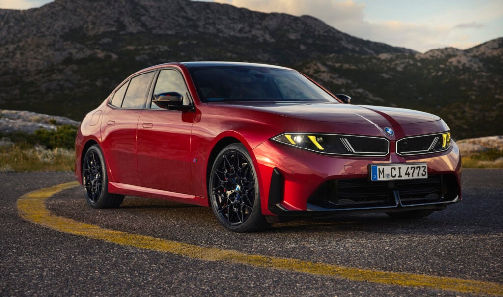Red BMW sedan in a three-quarter front view on a curved mountain road with rocky hills in the background.