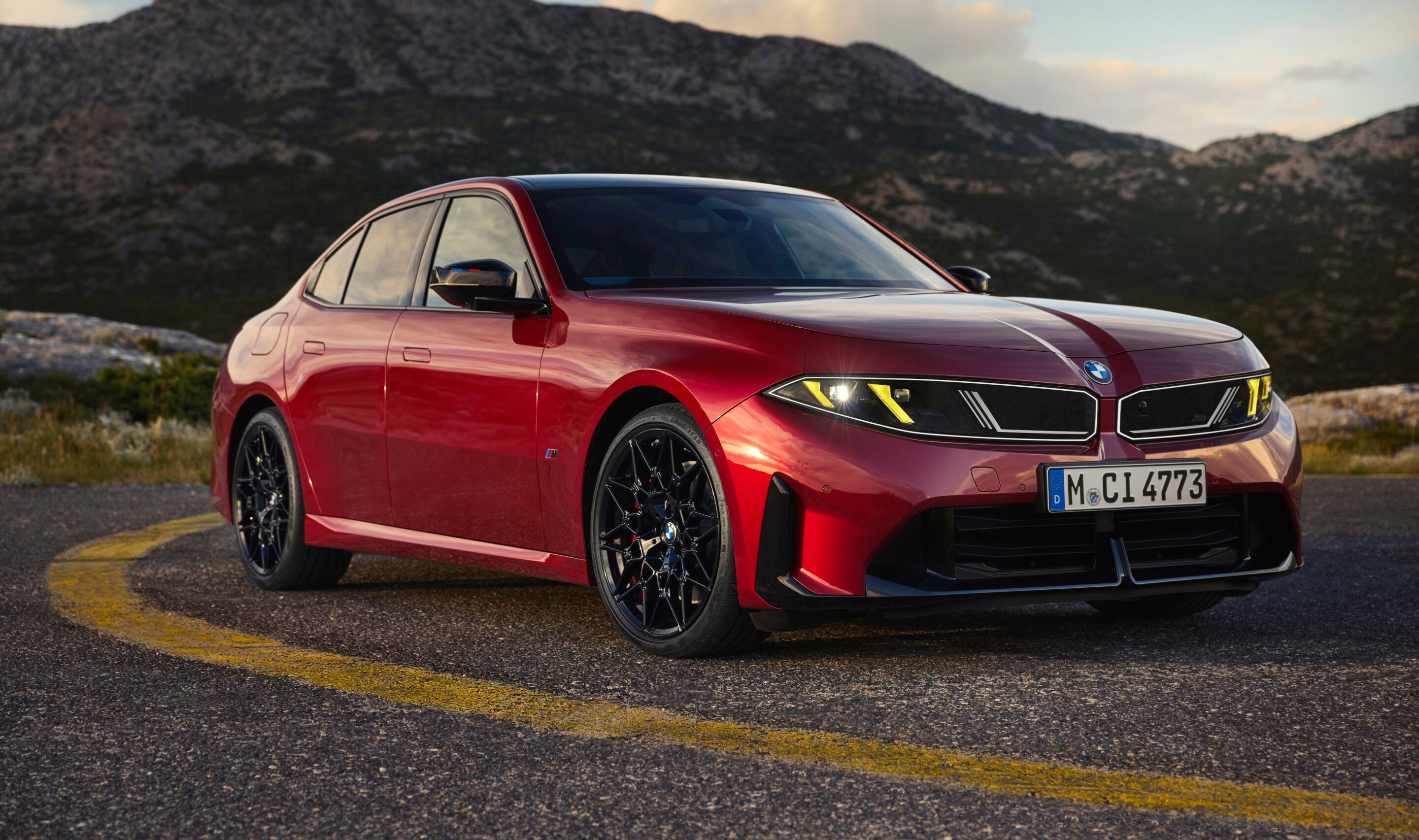 Red BMW sedan in a three-quarter front view on a curved mountain road with rocky hills in the background.