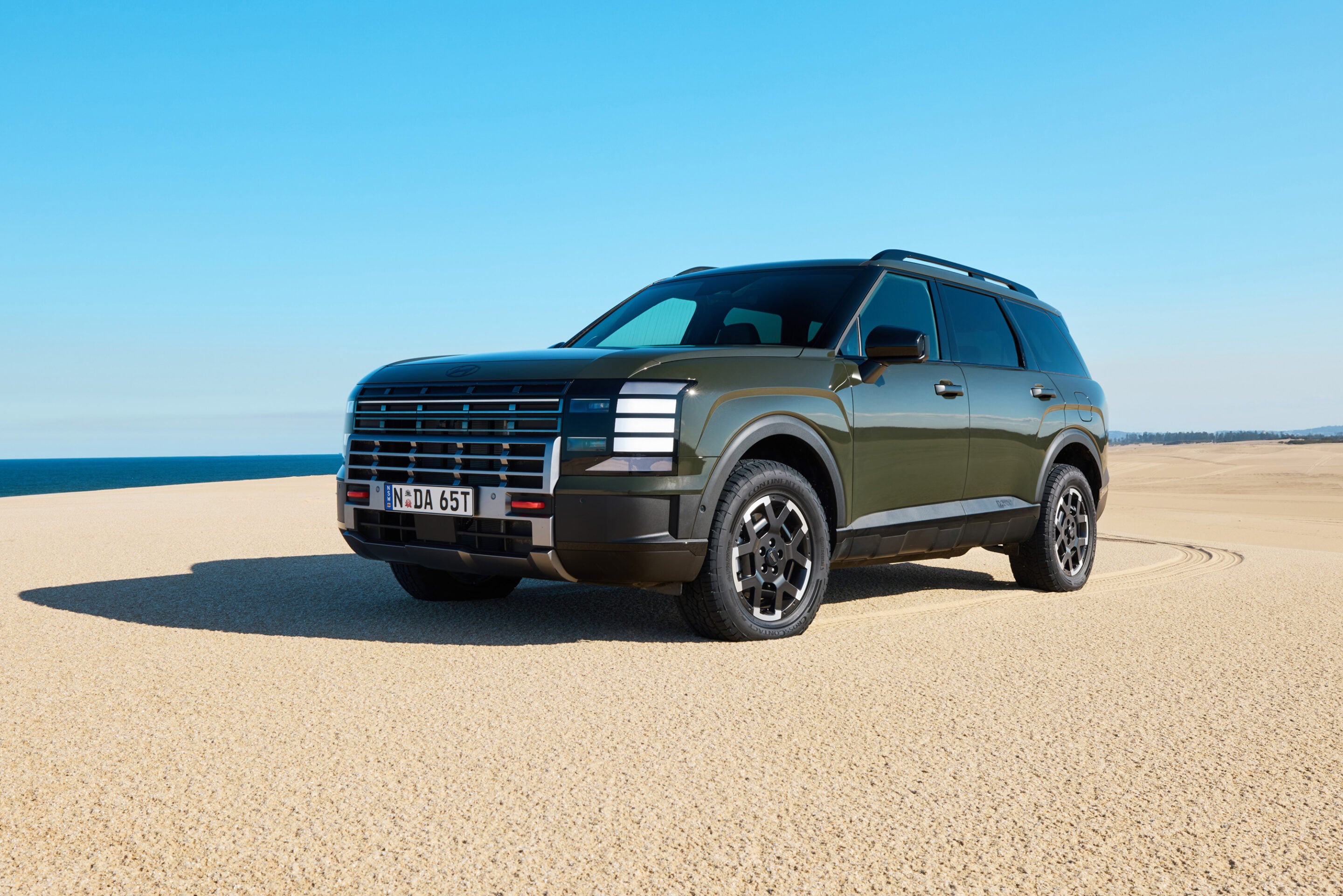 Front three-quarter view of a dark green SUV parked on a sandy beach with the blue ocean and sky in the background.