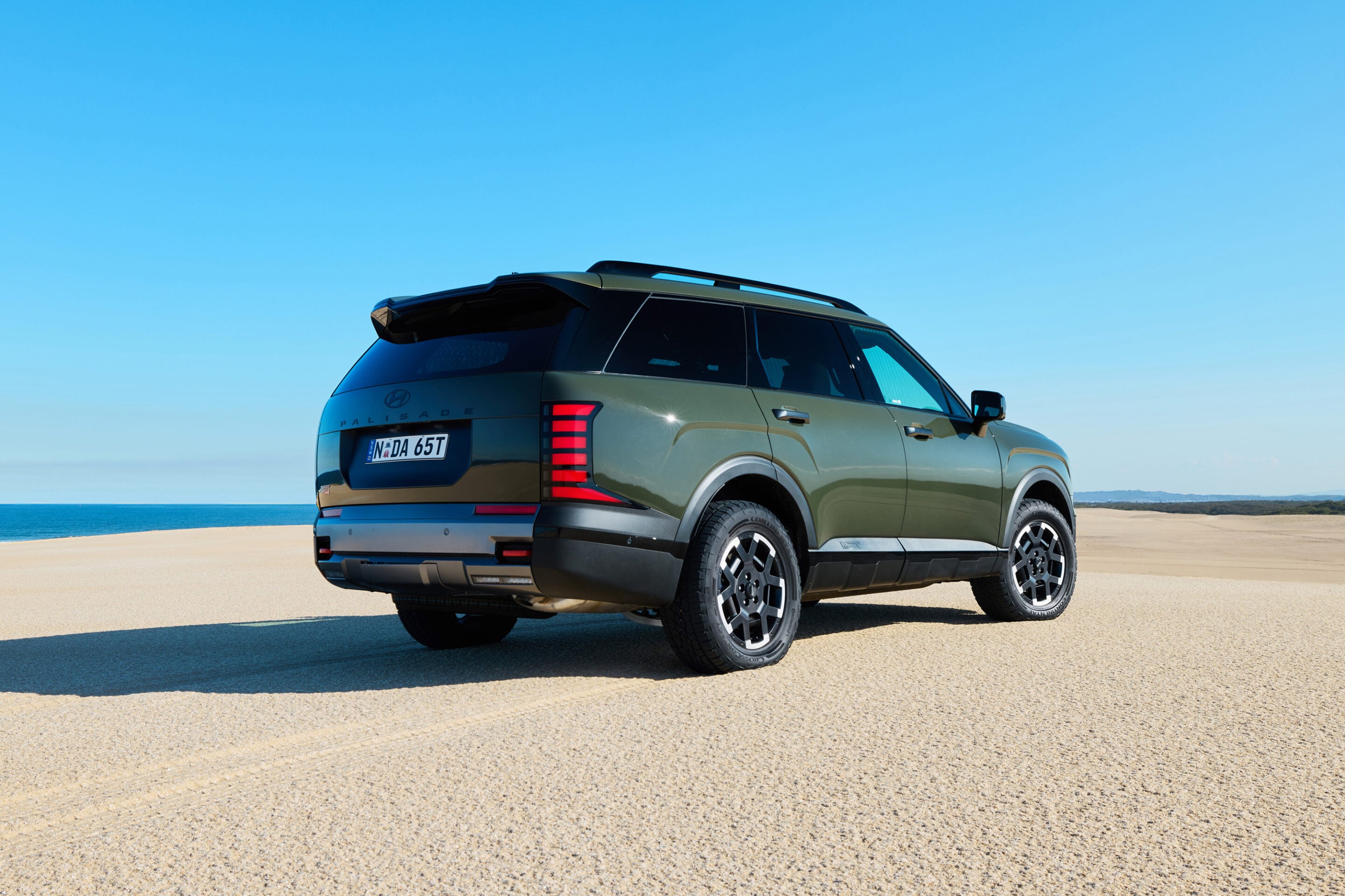 Green SUV parked on a sandy beach with a clear blue sky and ocean in the background.