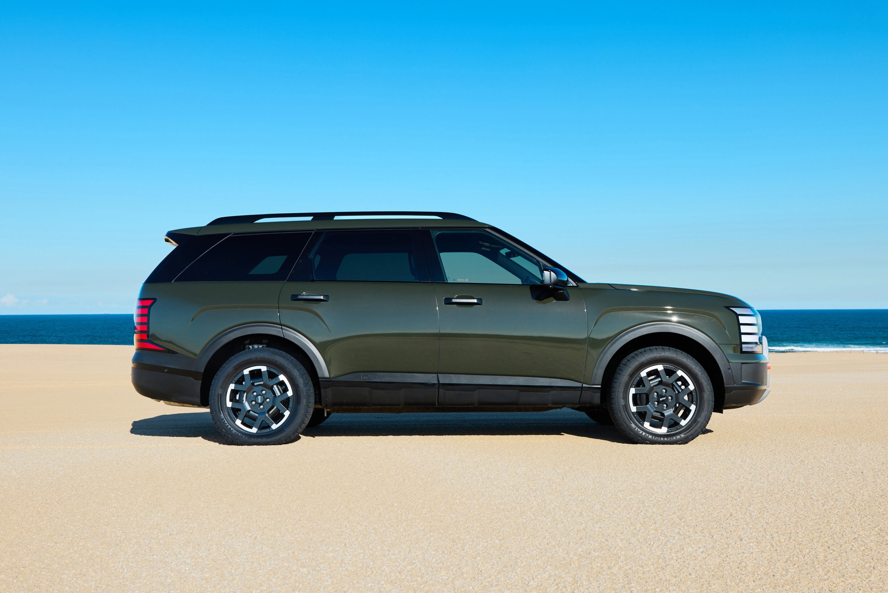 Green SUV parked on a sandy beach with a calm blue sea and bright blue sky in the background
