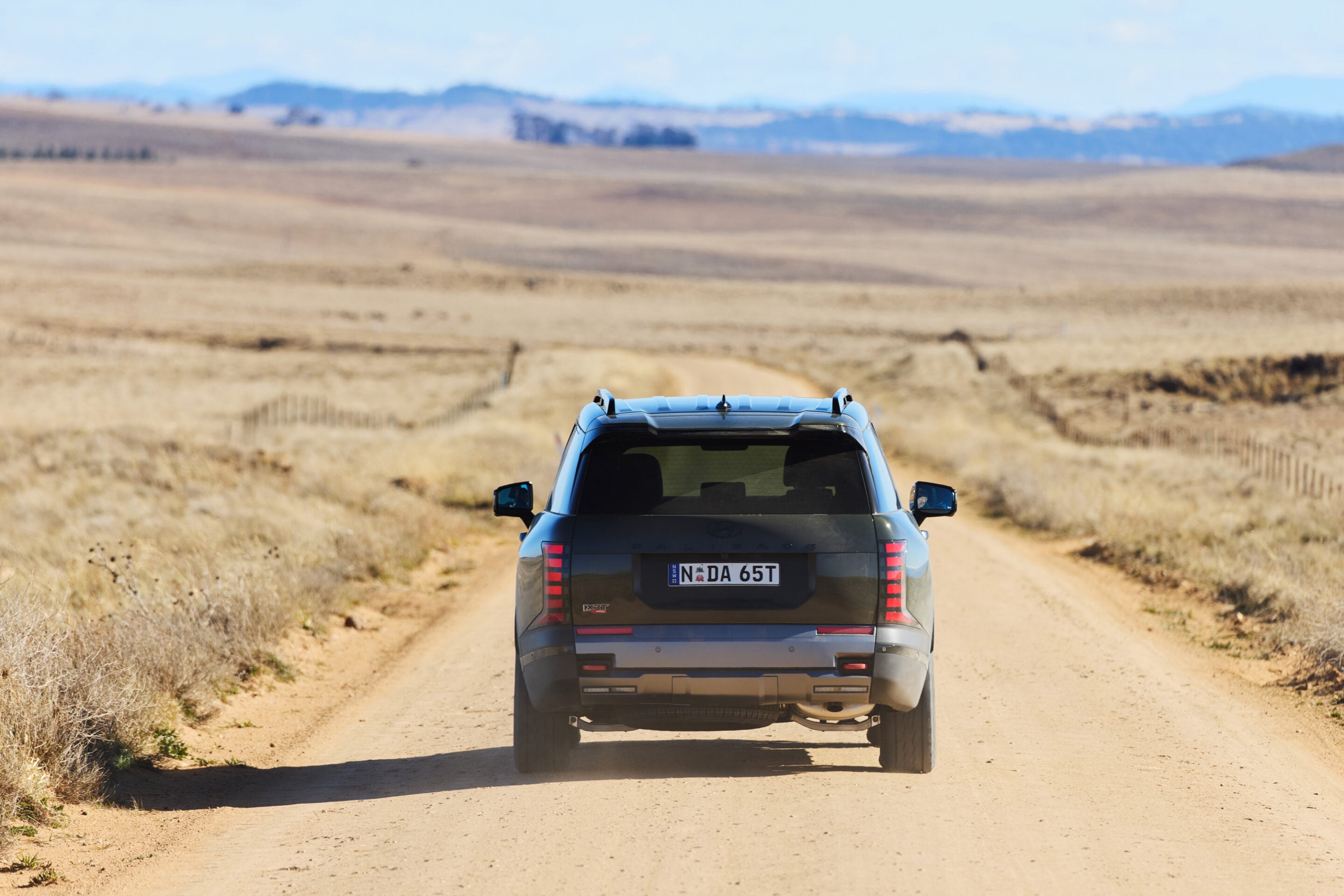 Rear view of a dark SUV driving down a dusty rural road through arid fields with hills on the horizon under a clear blue sky