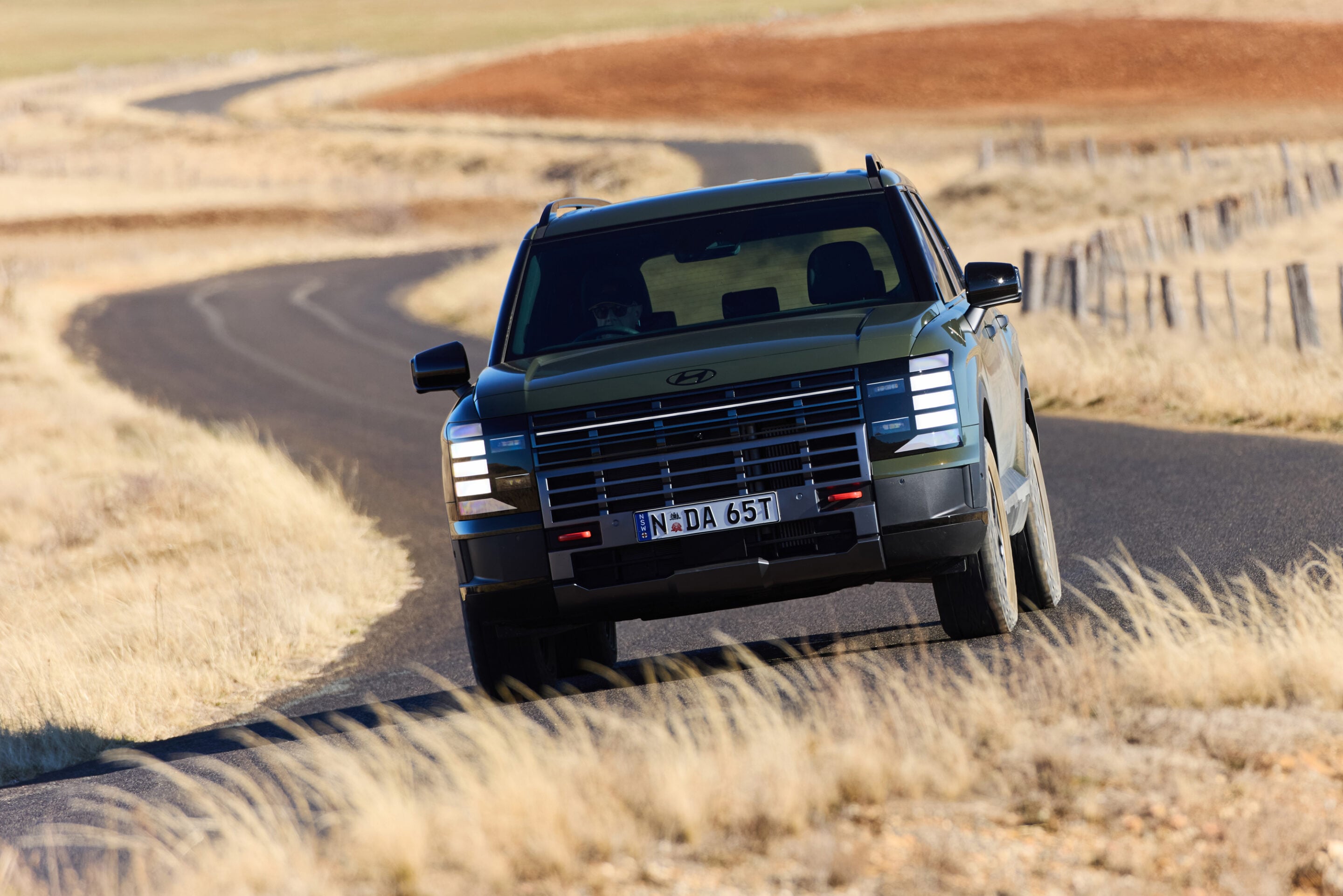Blue SUV driving on a curving rural road through a dry, grassy landscape; fence posts line the right side.