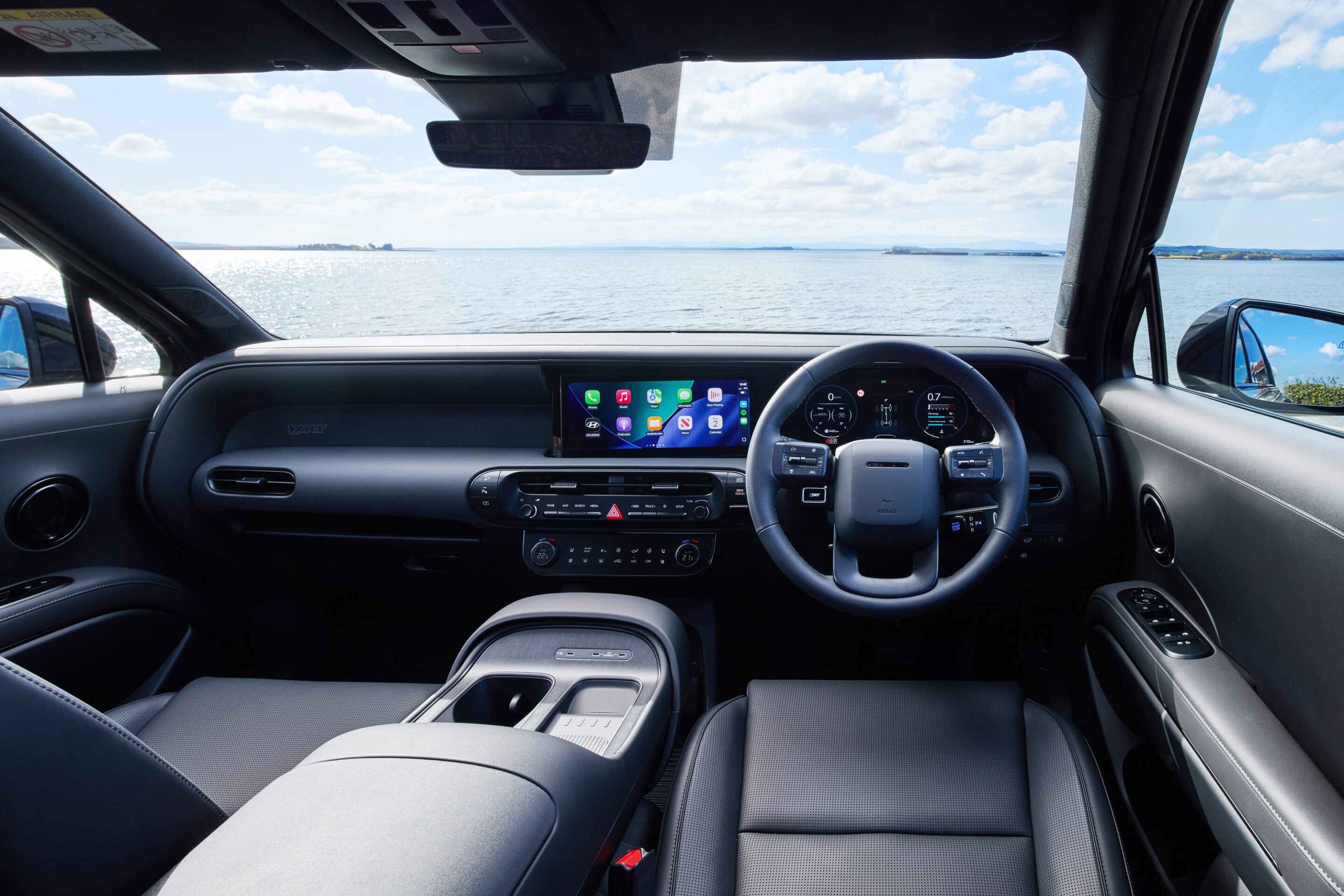 Interior of a car facing the ocean through the windshield, with the dashboard and touchscreen in view.