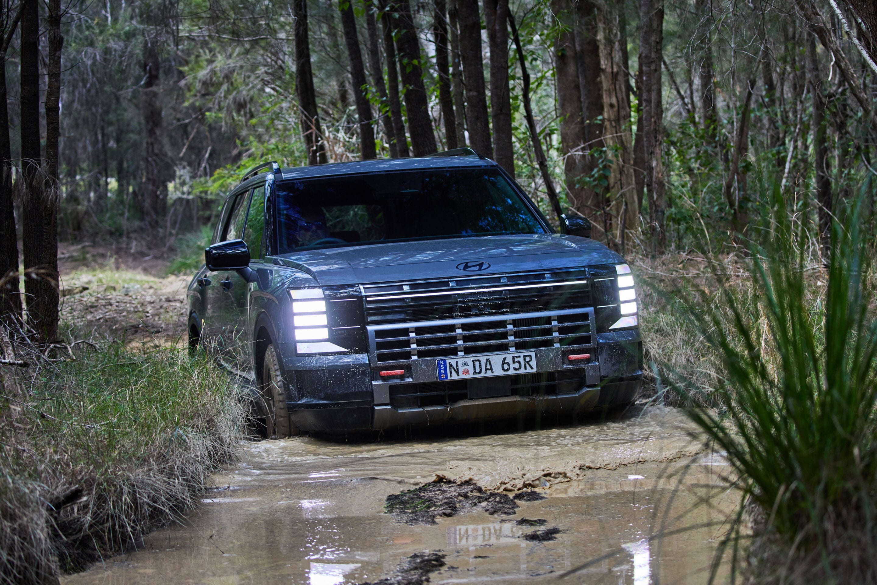 Dark SUV stuck on a muddy forest trail surrounded by trees and wet ground with water pooling around the tires.