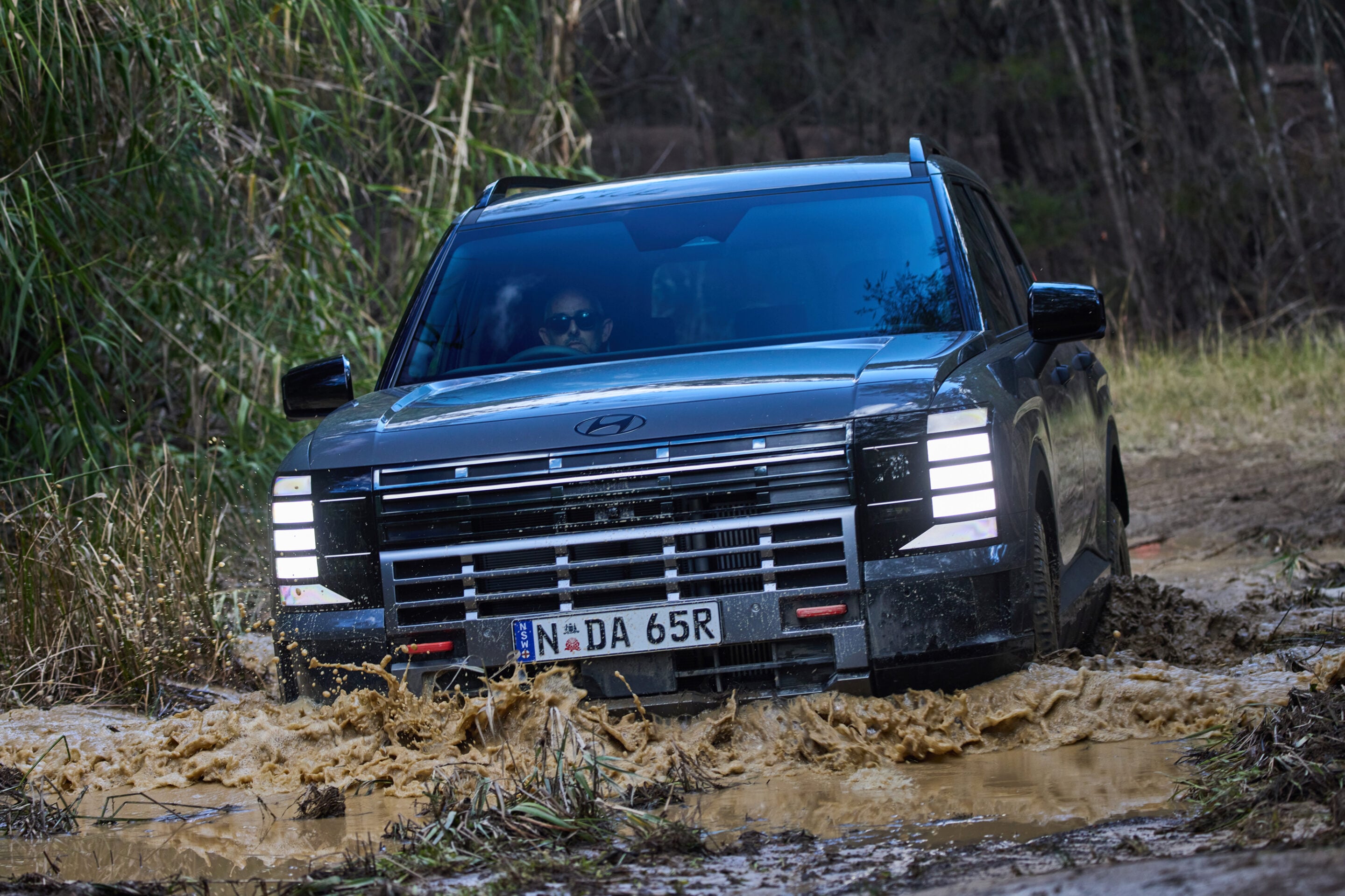 Blue SUV navigating a muddy, off-road path with splashing water near the wheels; driver wearing sunglasses visible through the windshield.