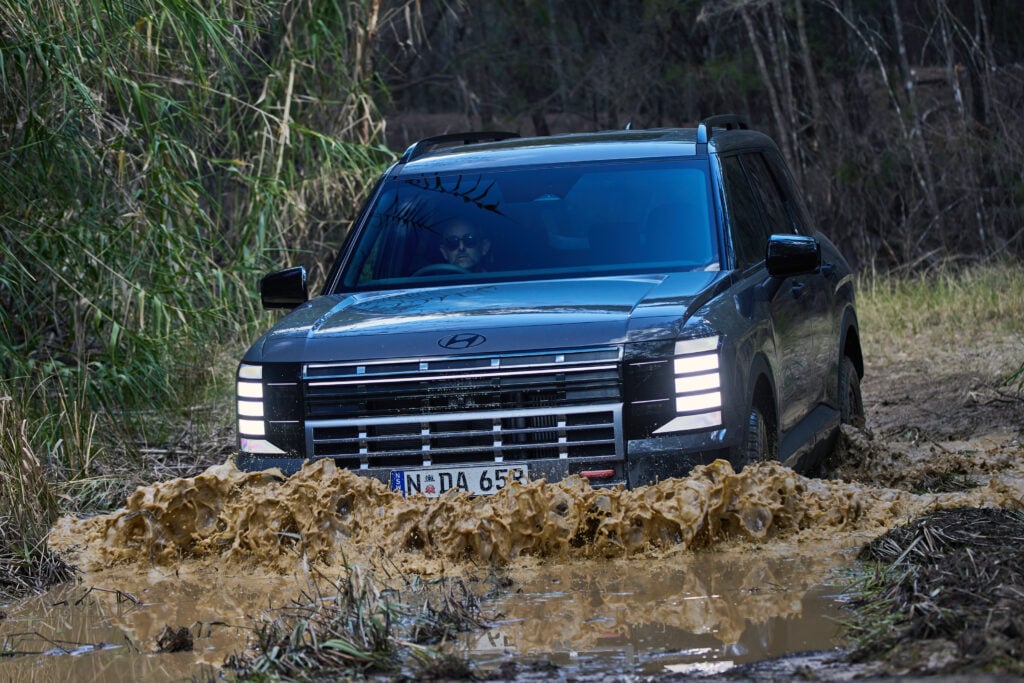 Front view of a blue SUV driving through a muddy puddle, spraying mud; driver wearing sunglasses visible inside the windshield.