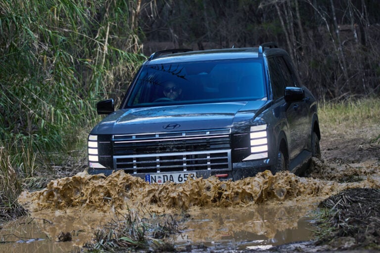 Front view of a blue SUV driving through a muddy puddle, spraying mud; driver wearing sunglasses visible inside the windshield.