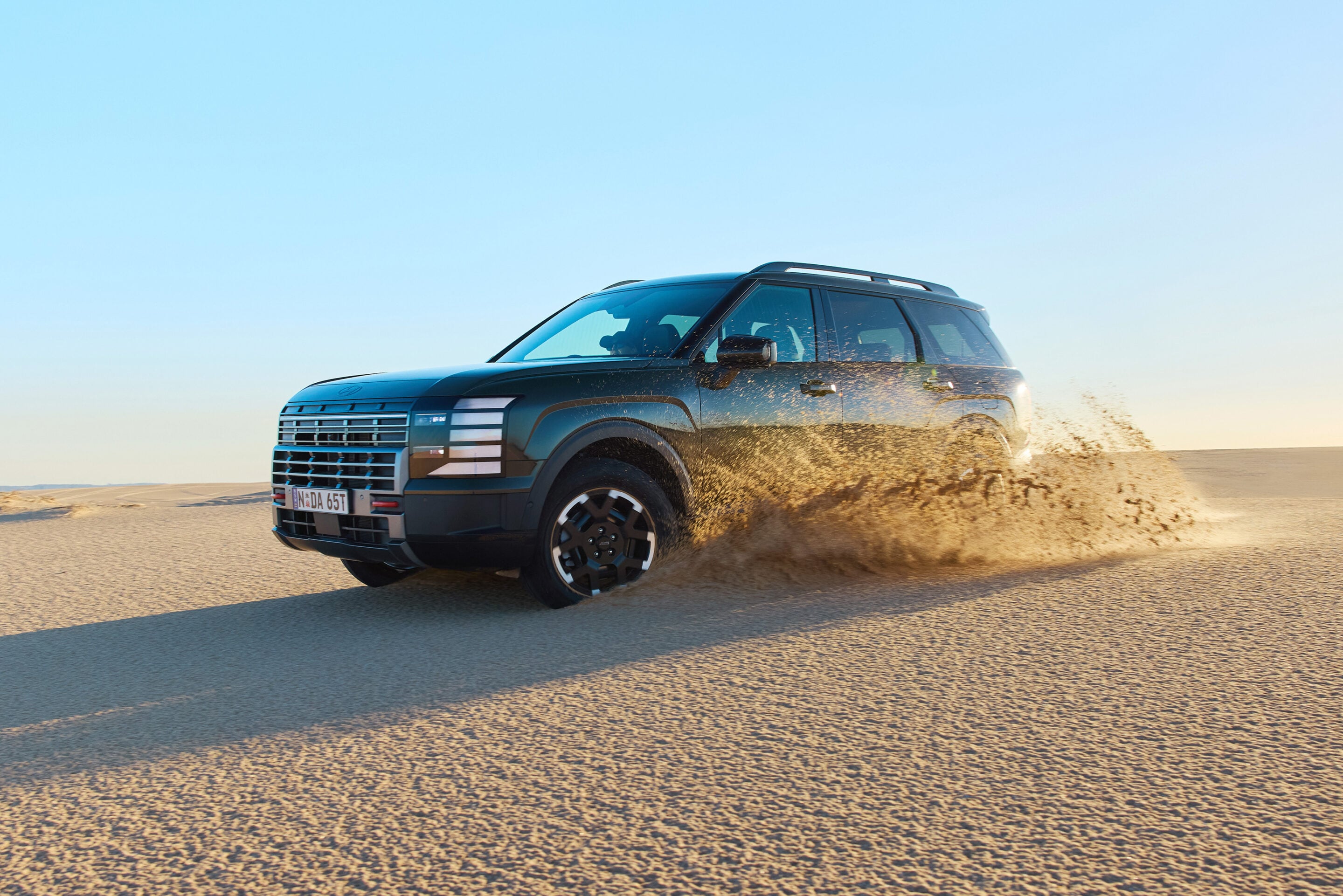 Dark blue SUV kicks up a plume of sand as it drives across a desert under a clear blue sky.
