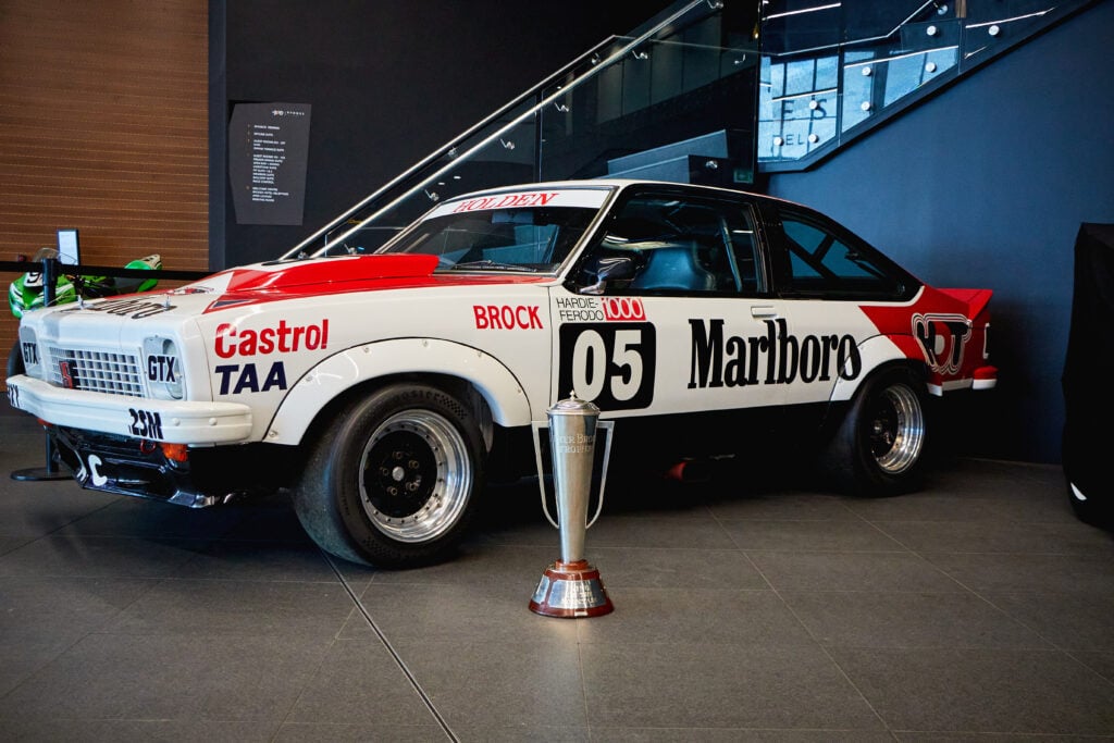 White race car on display with red Marlboro livery and number 05, positioned near a trophy in front of it.