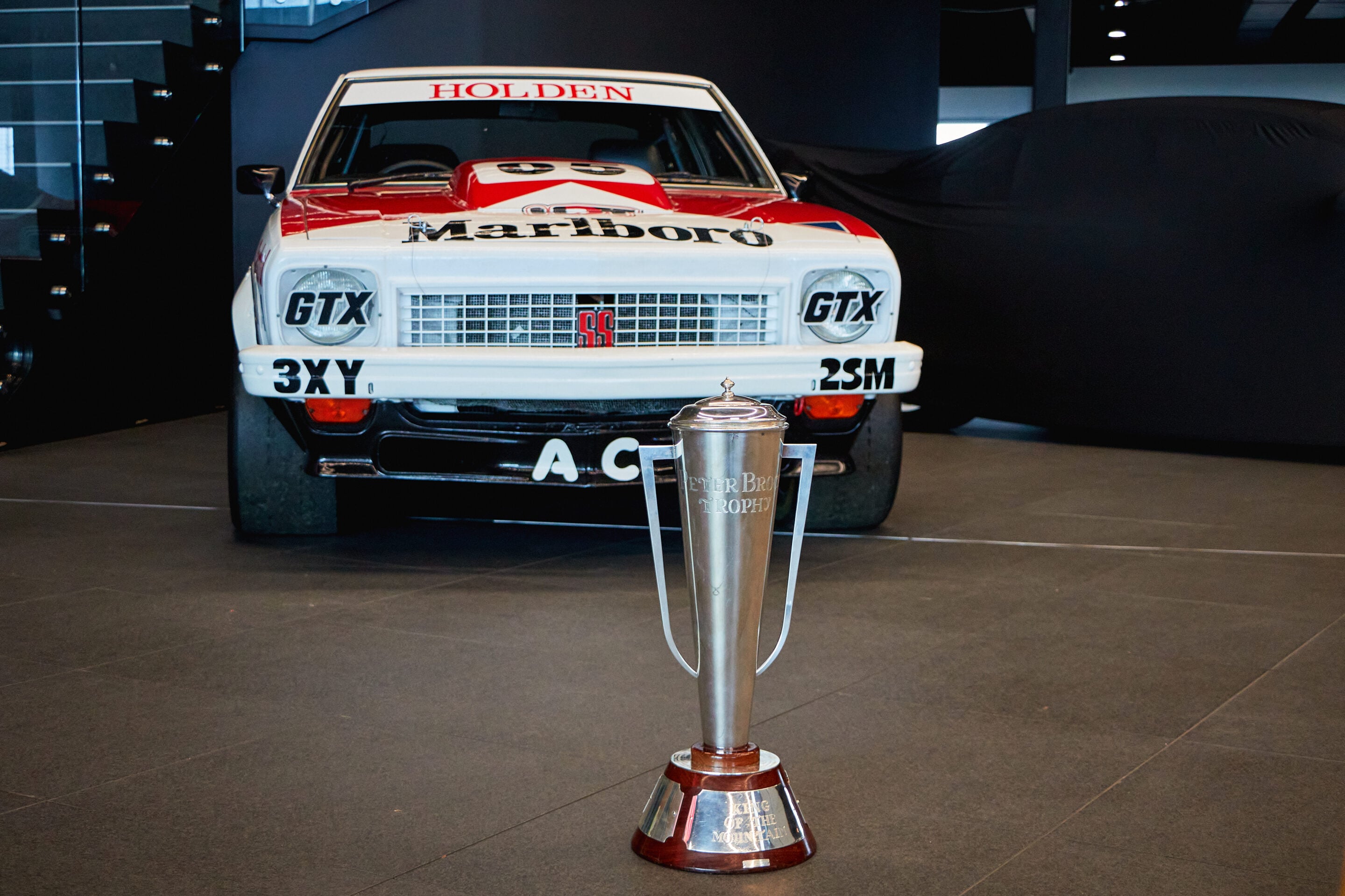 Winner's trophy set in front of a white and red classic Holden race car on display.