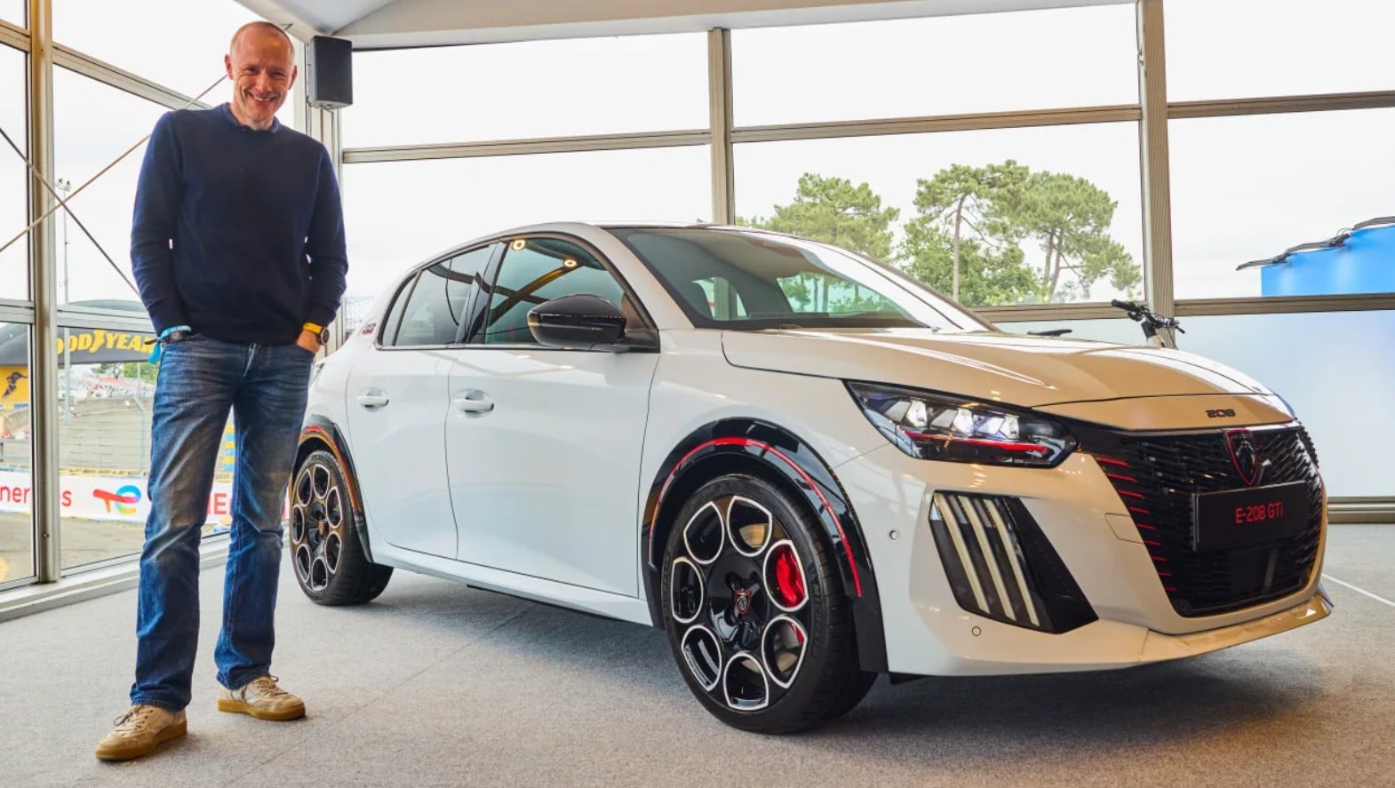 Smiling man in a navy sweater and jeans stands beside a white Peugeot E-208 GTi in a bright showroom.