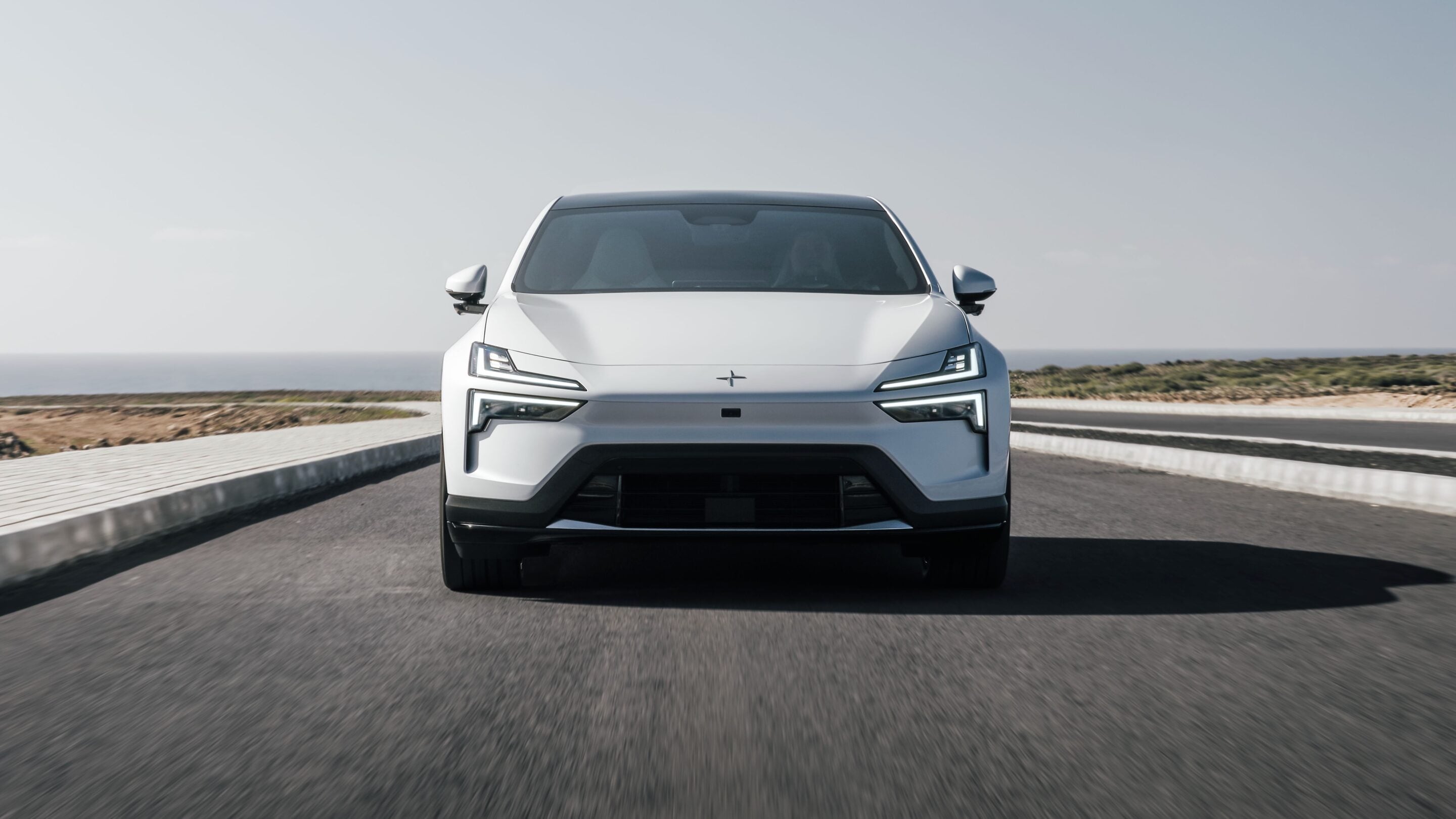 Front view of a white electric car driving toward the camera on a coastal highway with the sea in the background.