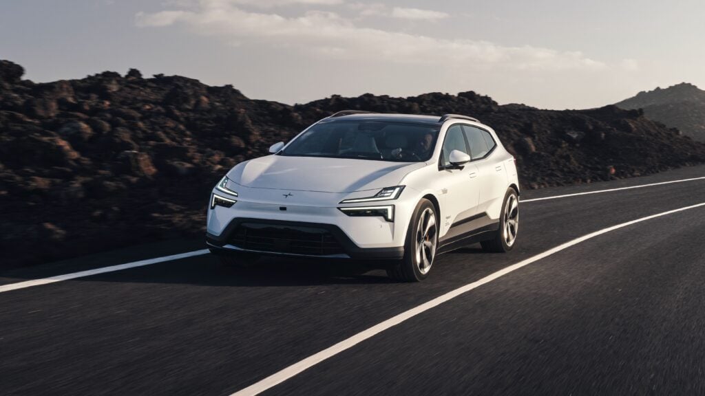 White futuristic SUV driving along a mountain road with rocky lava-like hills in the background, driver visible through the windshield.