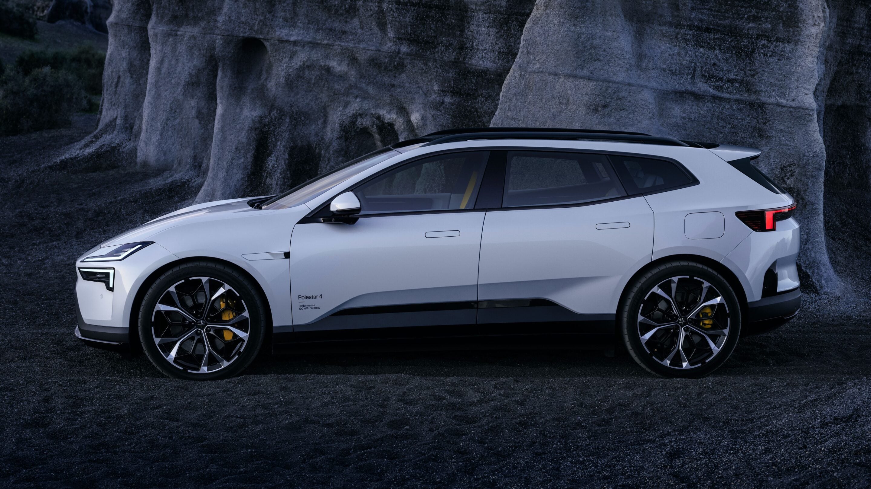 White Polestar 4 SUV parked on dark gravel with a rocky cliff backdrop, shown in a side profile at dusk.