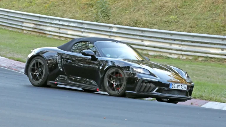 Black convertible sports car (Porsche Boxster) on a race track, leaning into a turn with a metal guardrail in the background.