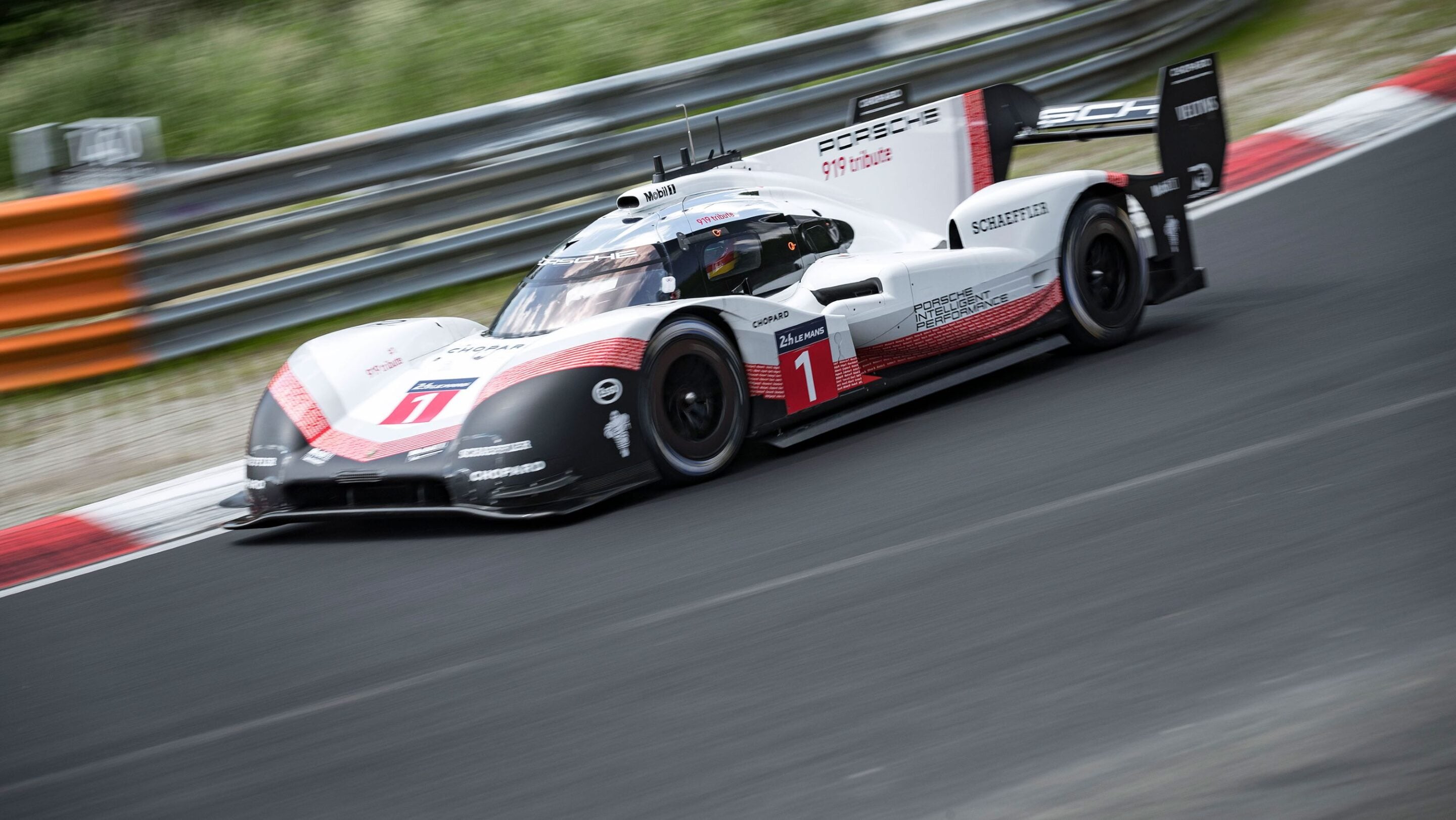White Porsche race car #1 speeding along a racetrack with safety barriers and greenery in the background, decals visible on the body.