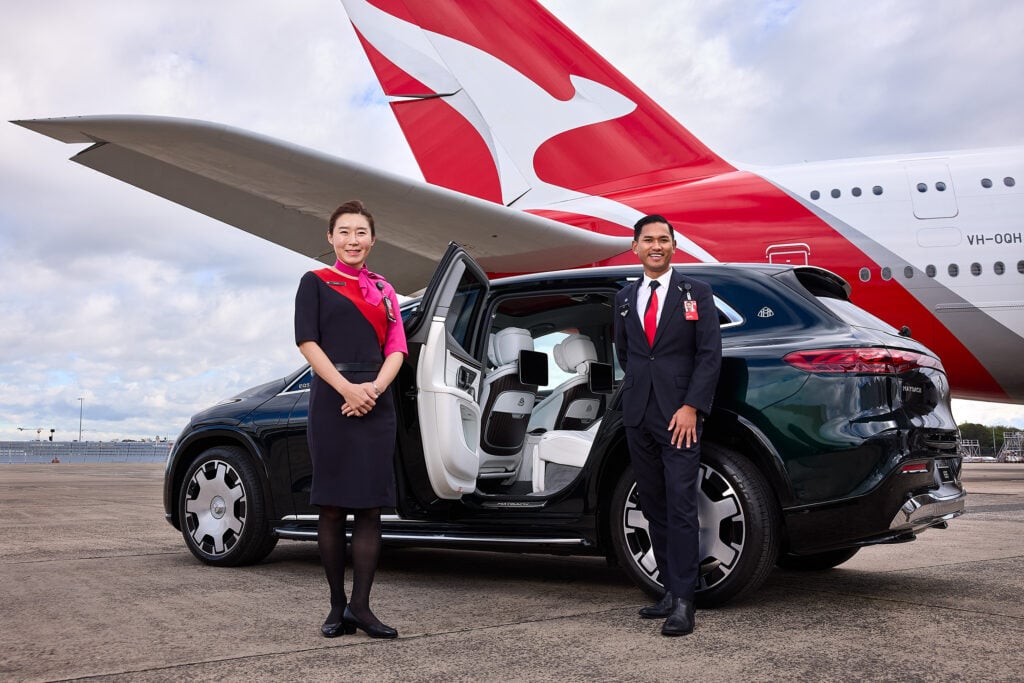Two airline staff members stand beside an open car on a tarmac with a red-and-white airplane tail in the background, smiling at the camera.