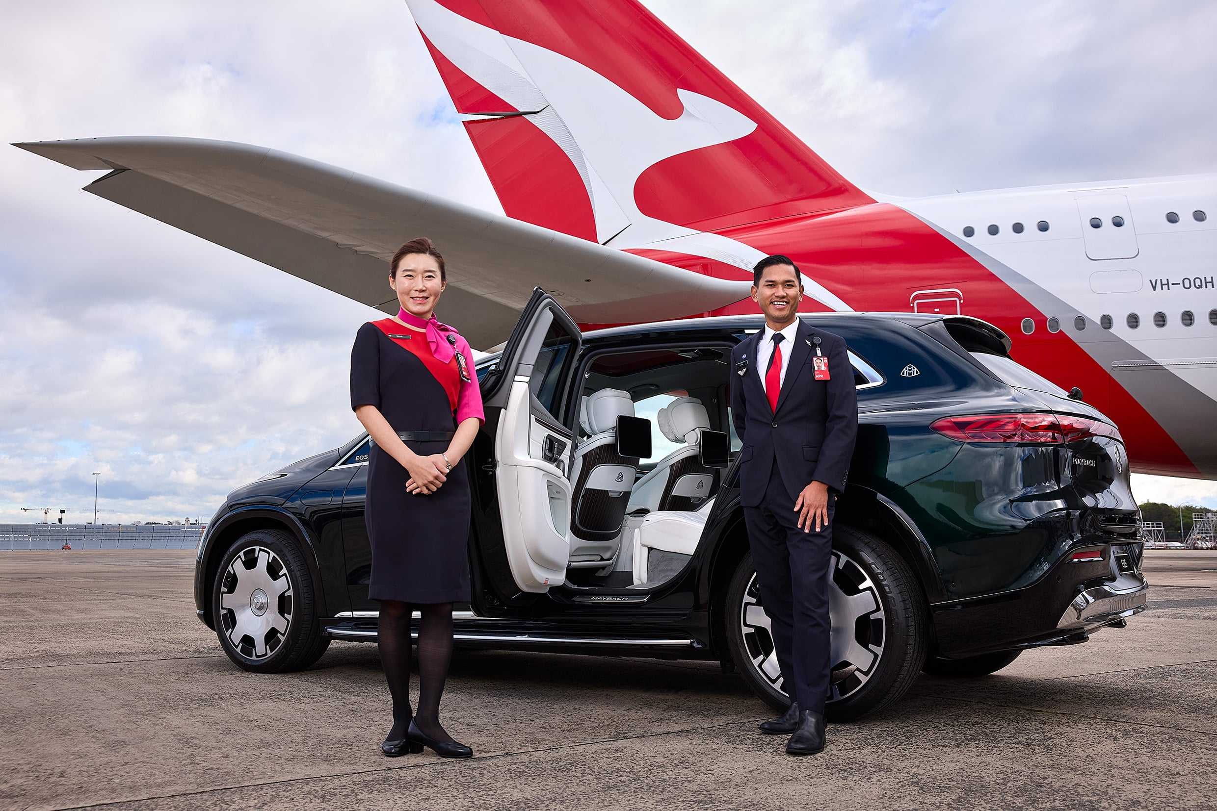 Two airline staff members stand beside an open car on a tarmac with a red-and-white airplane tail in the background, smiling at the camera.