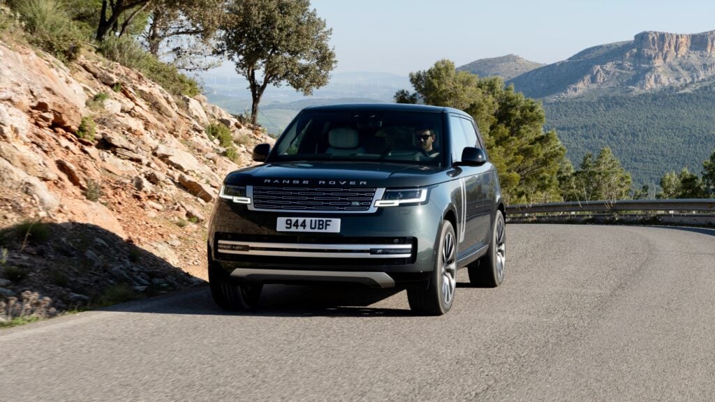 Range Rover SUV driving on a winding mountain road with a rocky cliff on the left and distant hills beyond the valley.