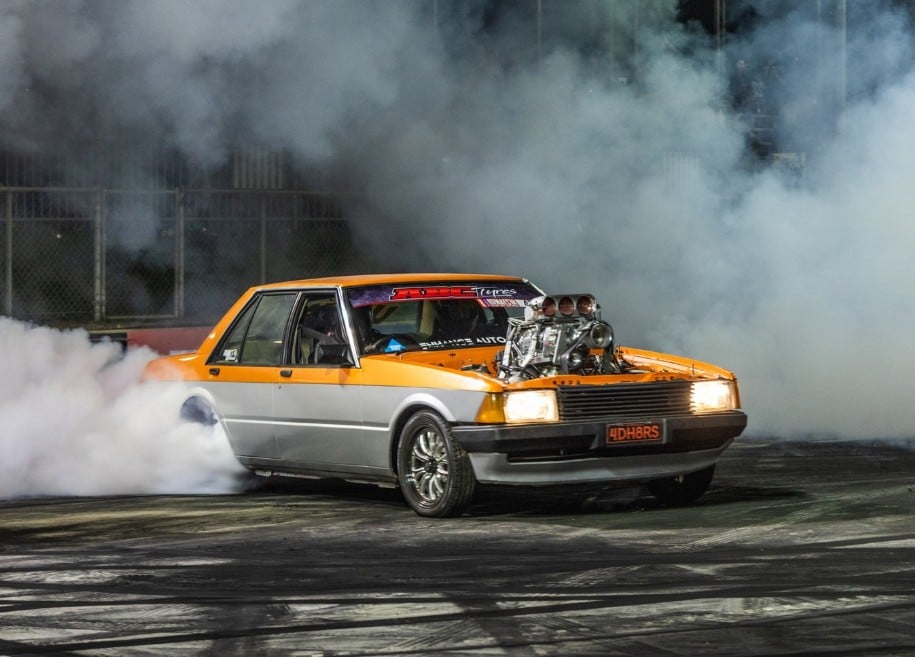 Orange and silver drag racing car performing a burnout, huge supercharged engine exposing plumes of smoke at a nighttime track.