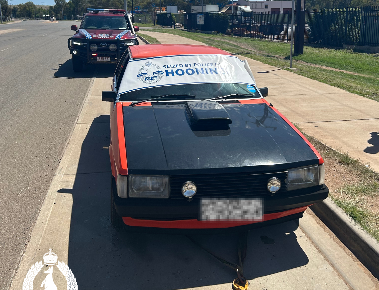 Red and black sedan with a banner reading 'Seized by Police HOONING' on the windshield; police SUV in pursuit behind on a sunny street surface of a road.