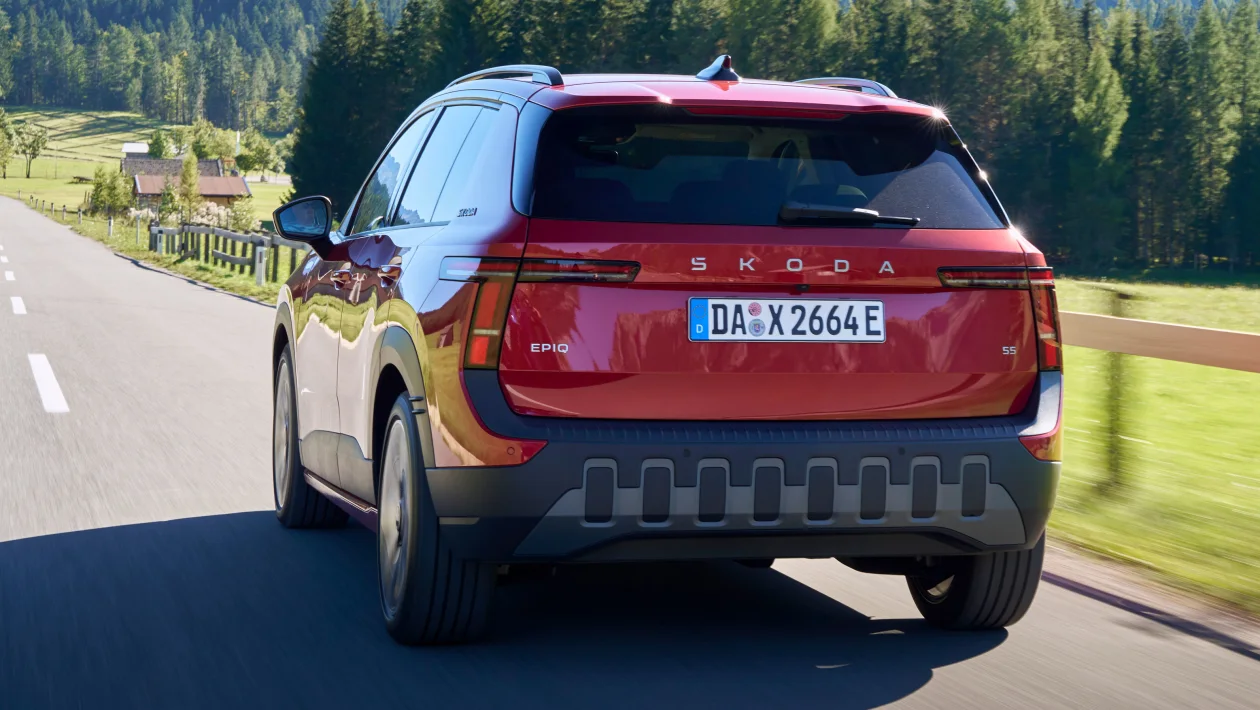 Rear view of a red Skoda Kodiaq SUV driving on a rural highway with green fields and forest in the background, license plate visible DA X 2664 E