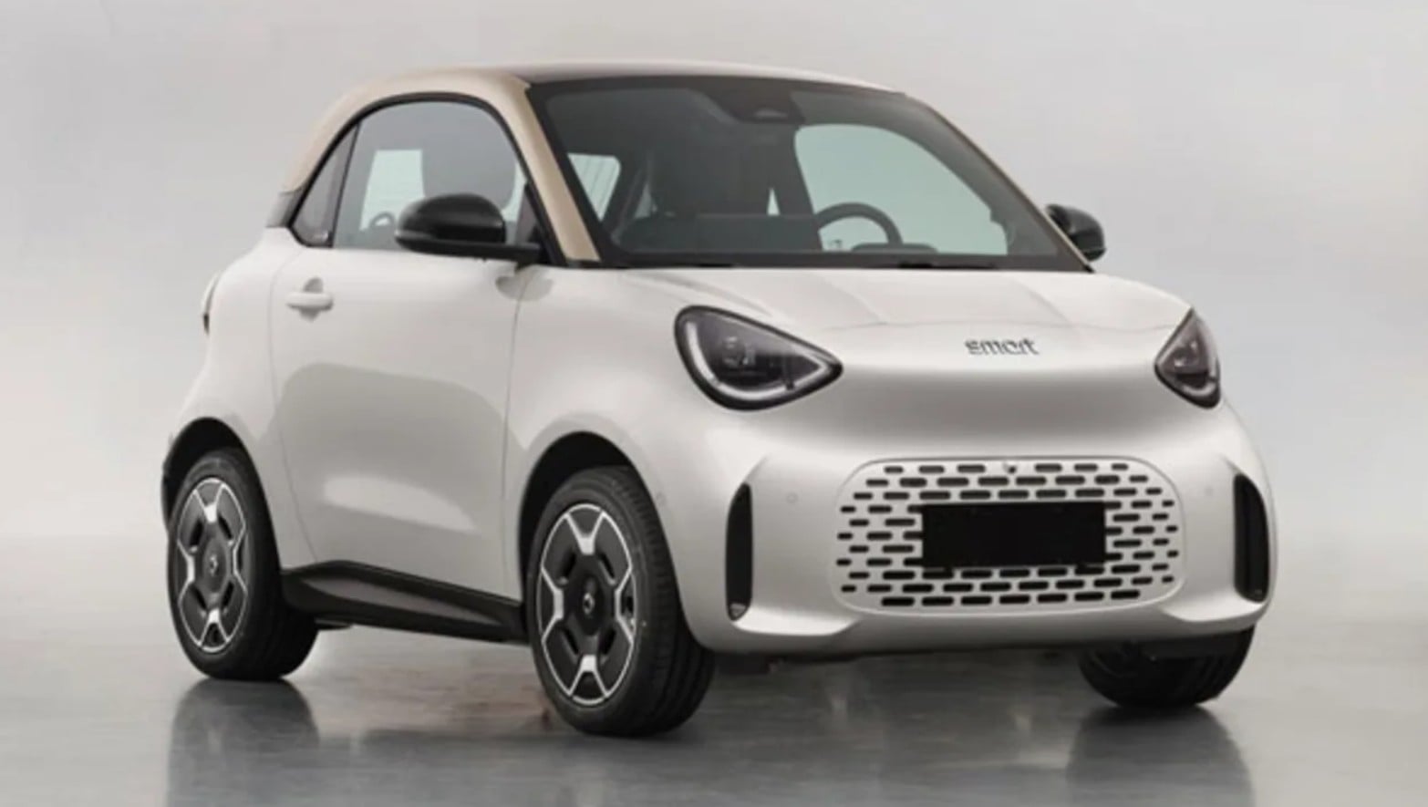 Three-quarter front view of a small white city car with a tan roof, indoors, showing its rounded headlights and perforated grille.