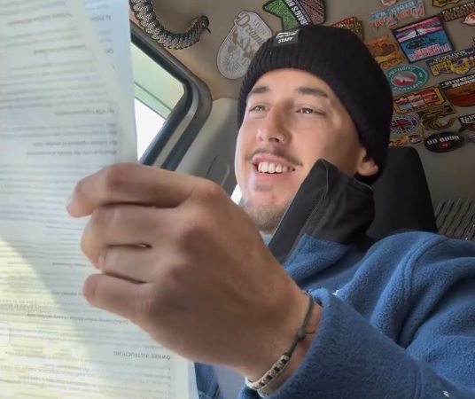 Young man in a beanie reading a document inside a sticker-covered van, smiling.