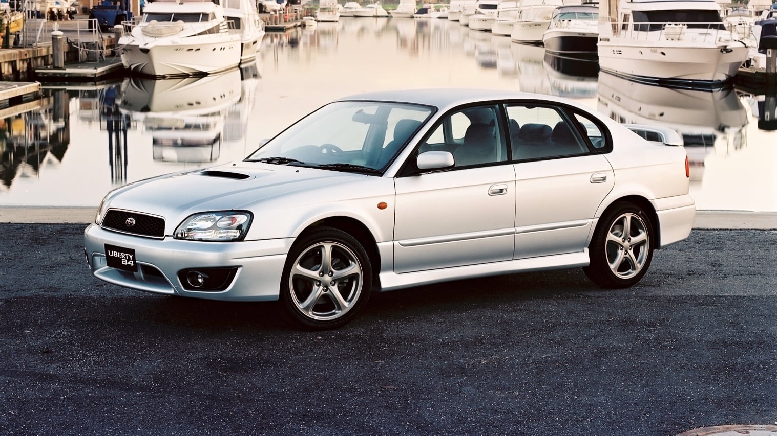 Silver sedan parked on an asphalt promenade by a marina with moored boats and calm water behind it, license plate reads LIBERTY B4.