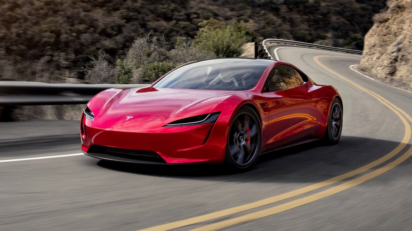 Red Tesla sports car driving along a winding mountain road with guardrails and blurred scenery