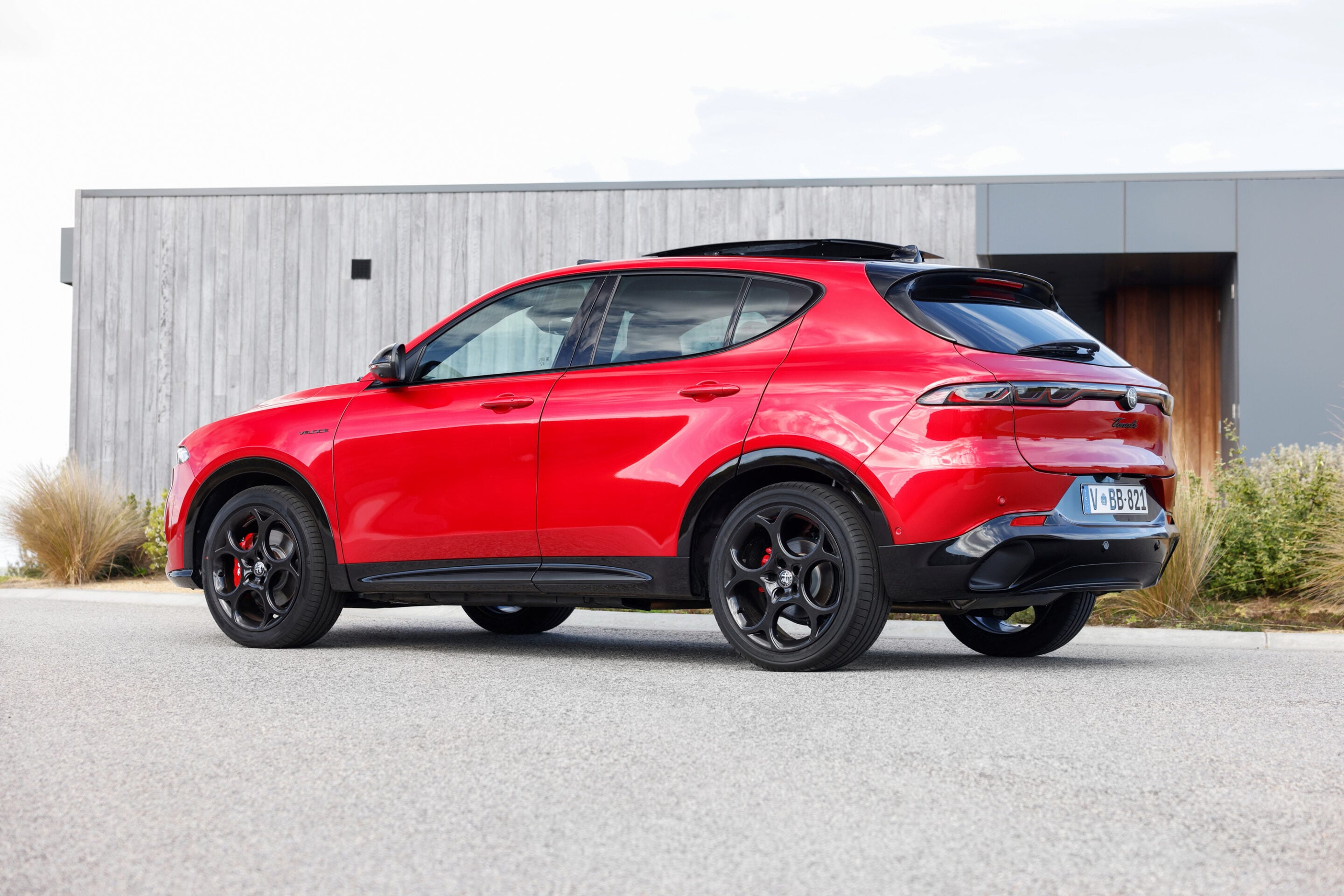 Red luxury SUV parked on a driveway beside a modern building with wooden panels; black wheels and roof rails visible in profile.