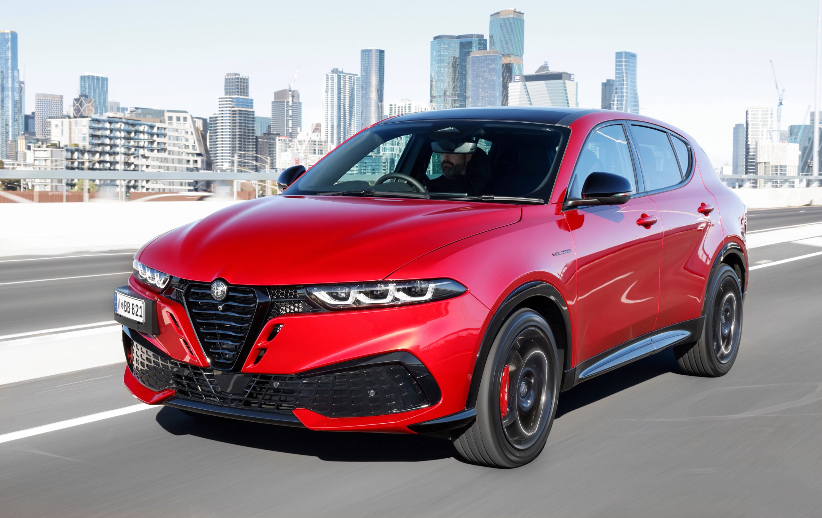 Red Alfa Romeo SUV driving on a highway with a modern city skyline in the background, motion blur suggesting speed.