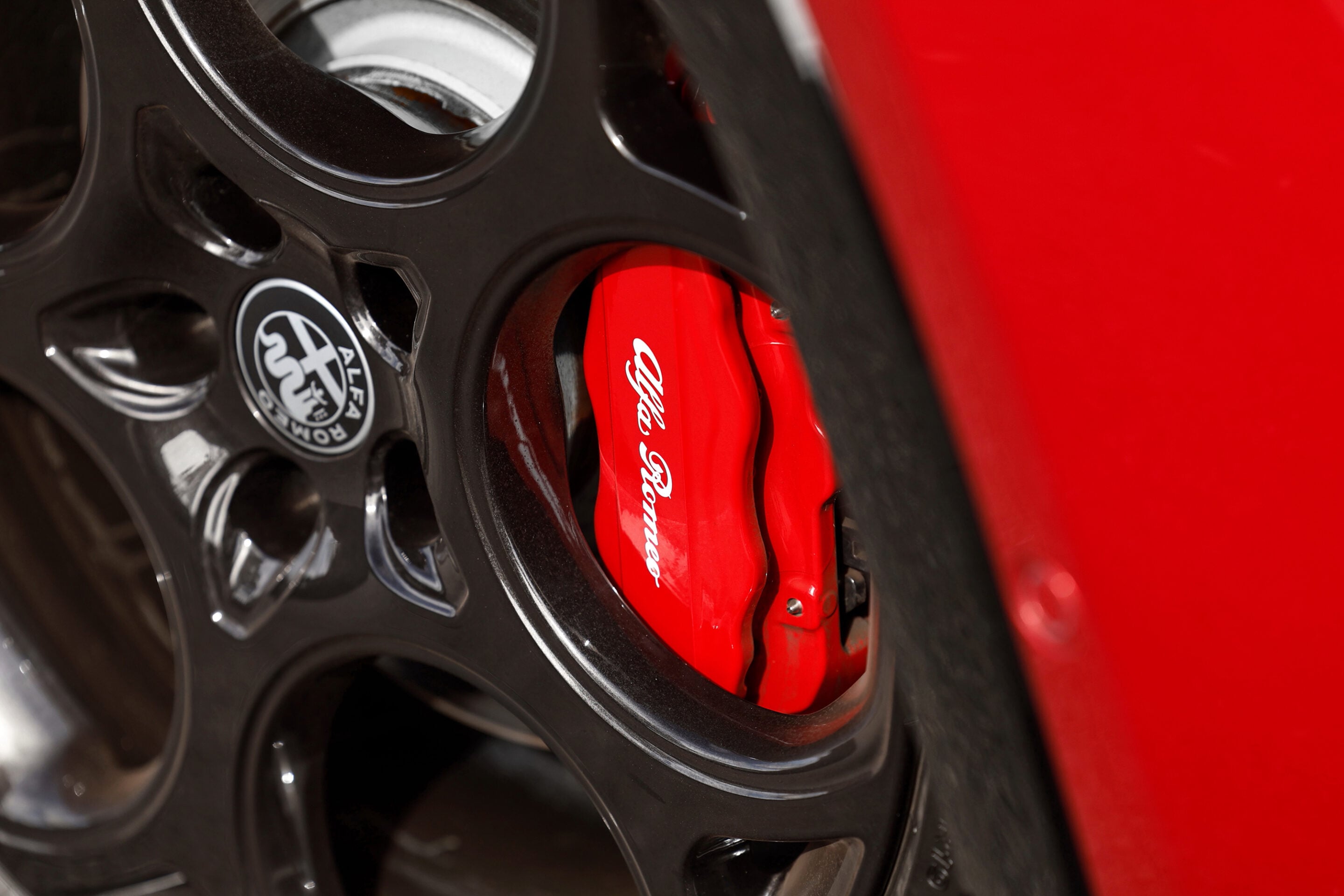 Close-up of a glossy red brake caliper with white Ferrari lettering, visible behind a black alloy wheel rim.