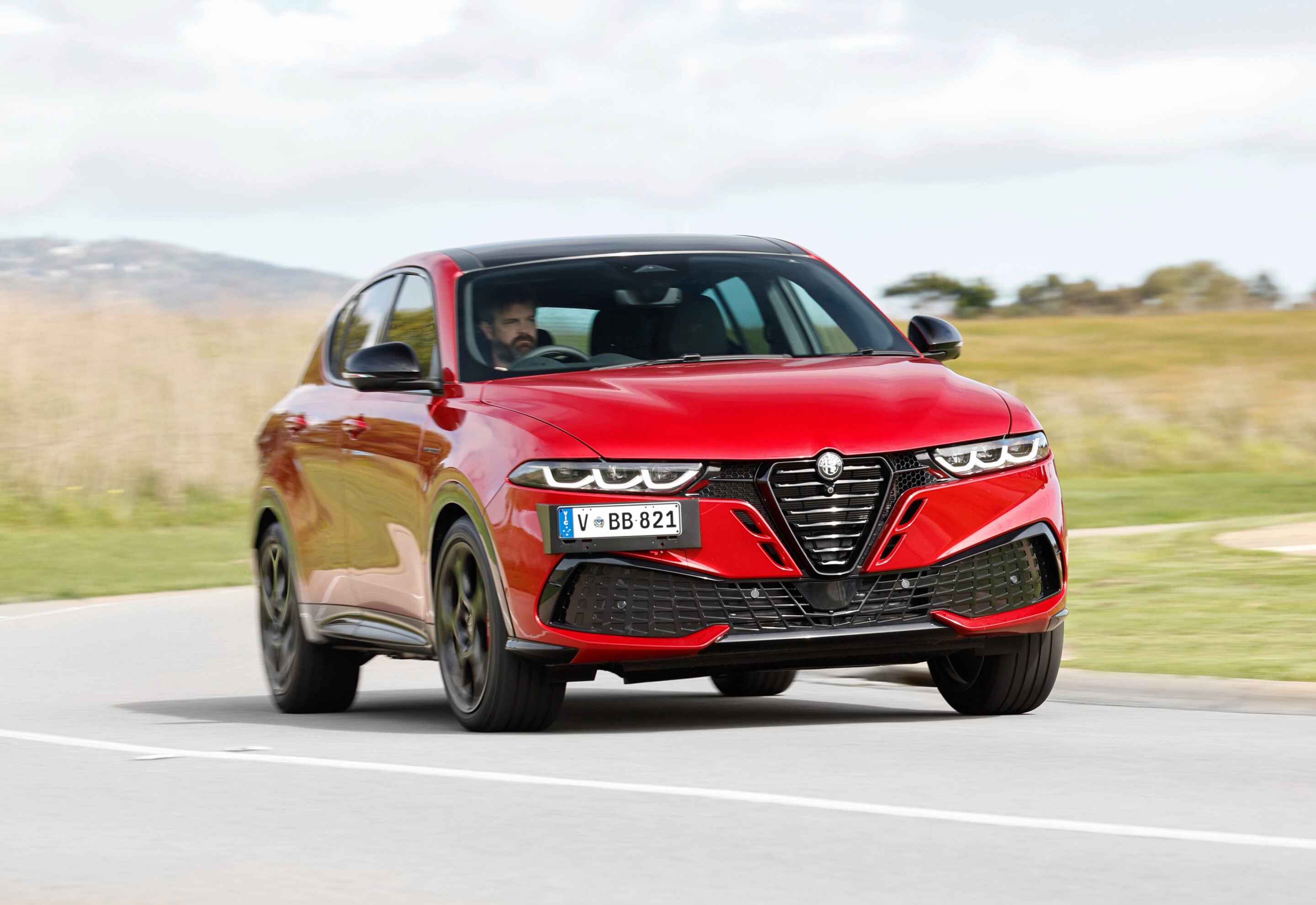 Red SUV driving along a rural highway, front-side view with motion blur on the wheels and grassy fields in the background, clear sky above.