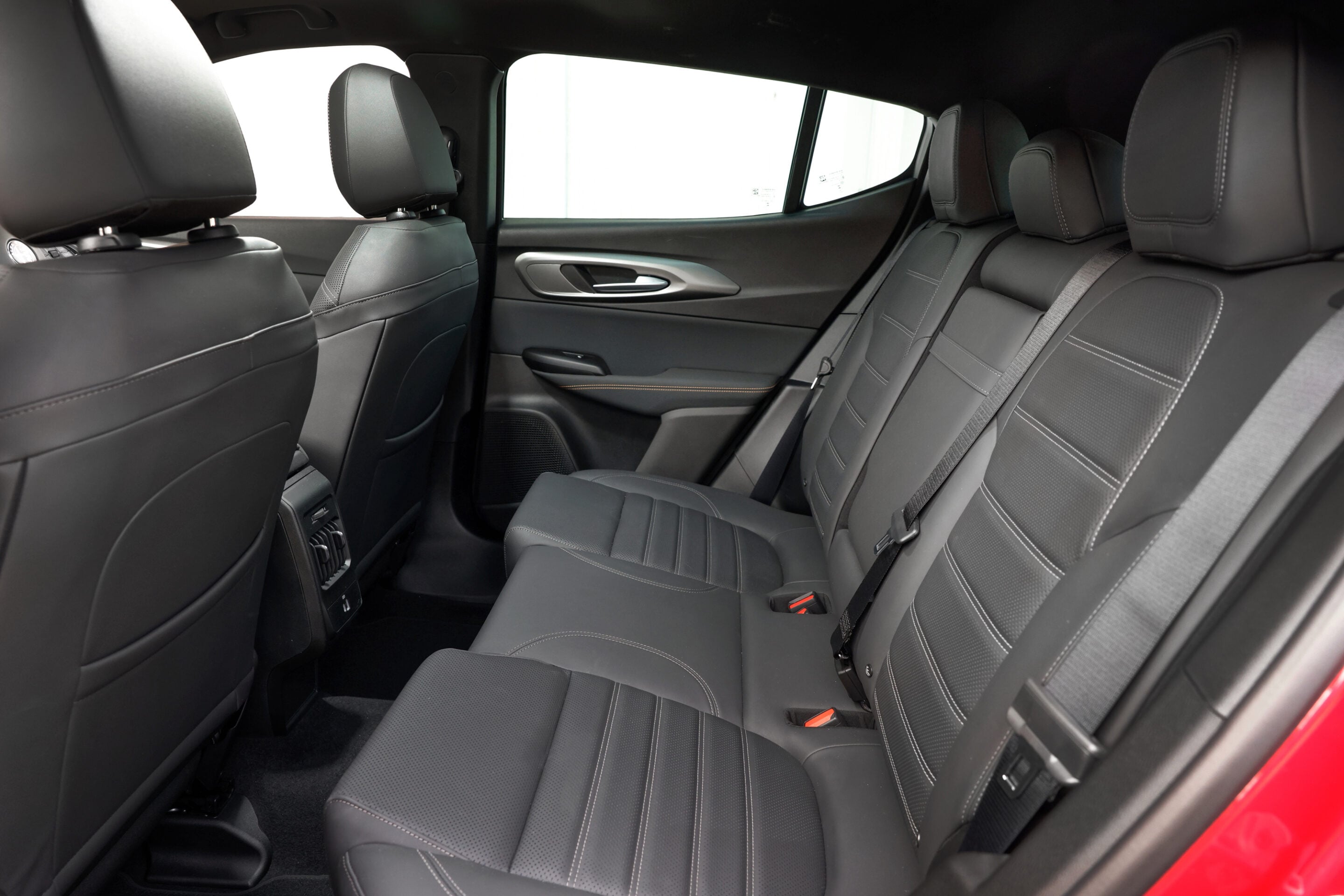 Back seat area of a modern car with black leather seats and red exterior visible at the door edge, with headrests and seat belts visible.