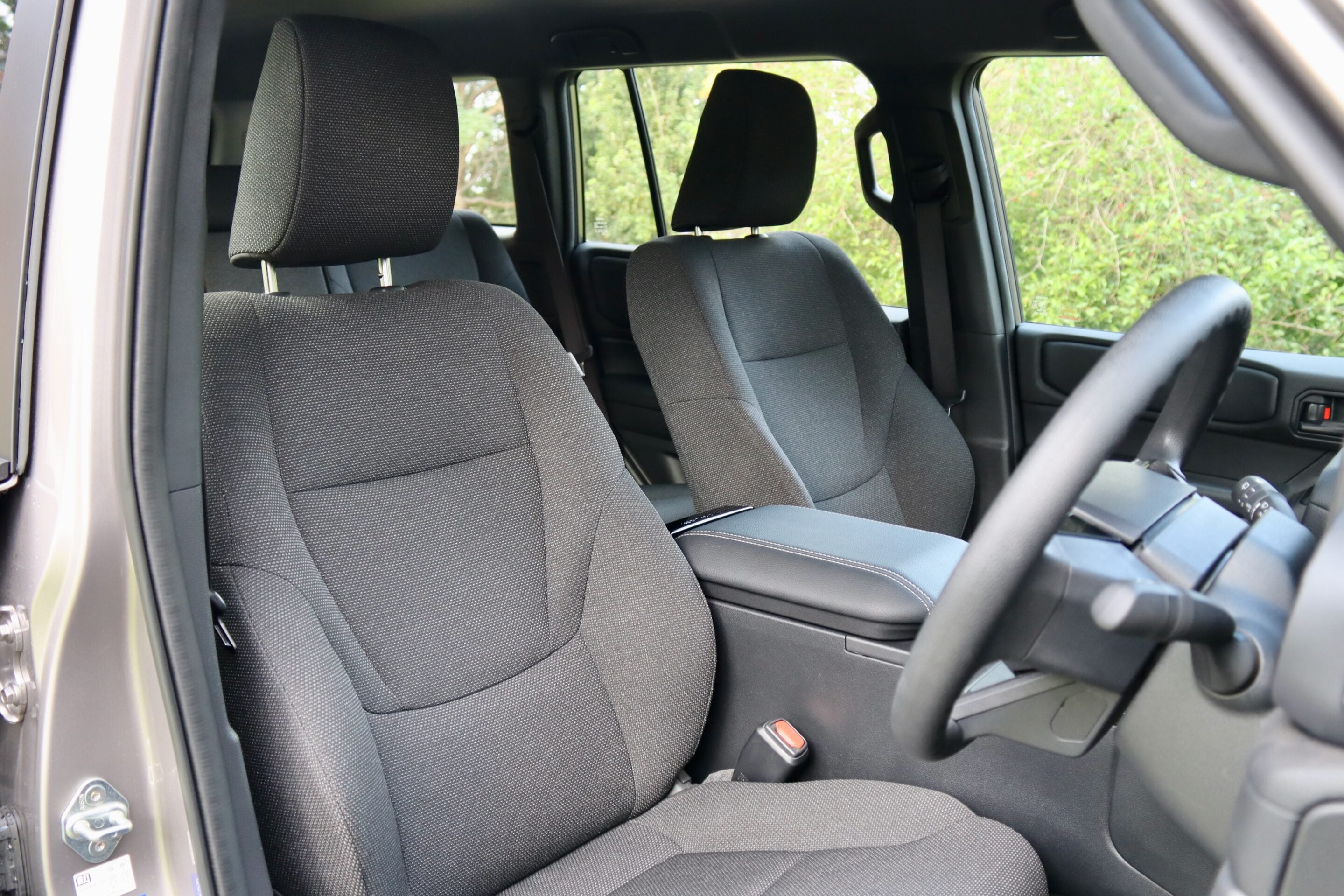 Interior of a car showing two front black fabric seats with headrests and a steering wheel visible on the right side, greenery outside the window.