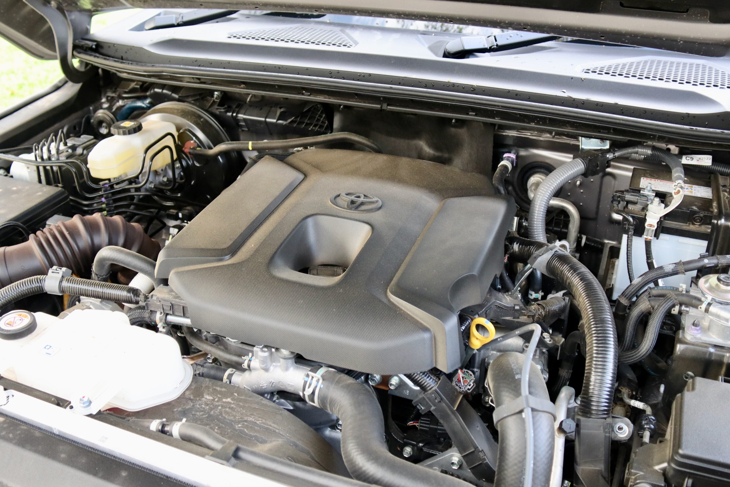 Front half of the car's engine bay with a black plastic engine cover bearing a Toyota emblem, plus hoses and wiring surrounding it.