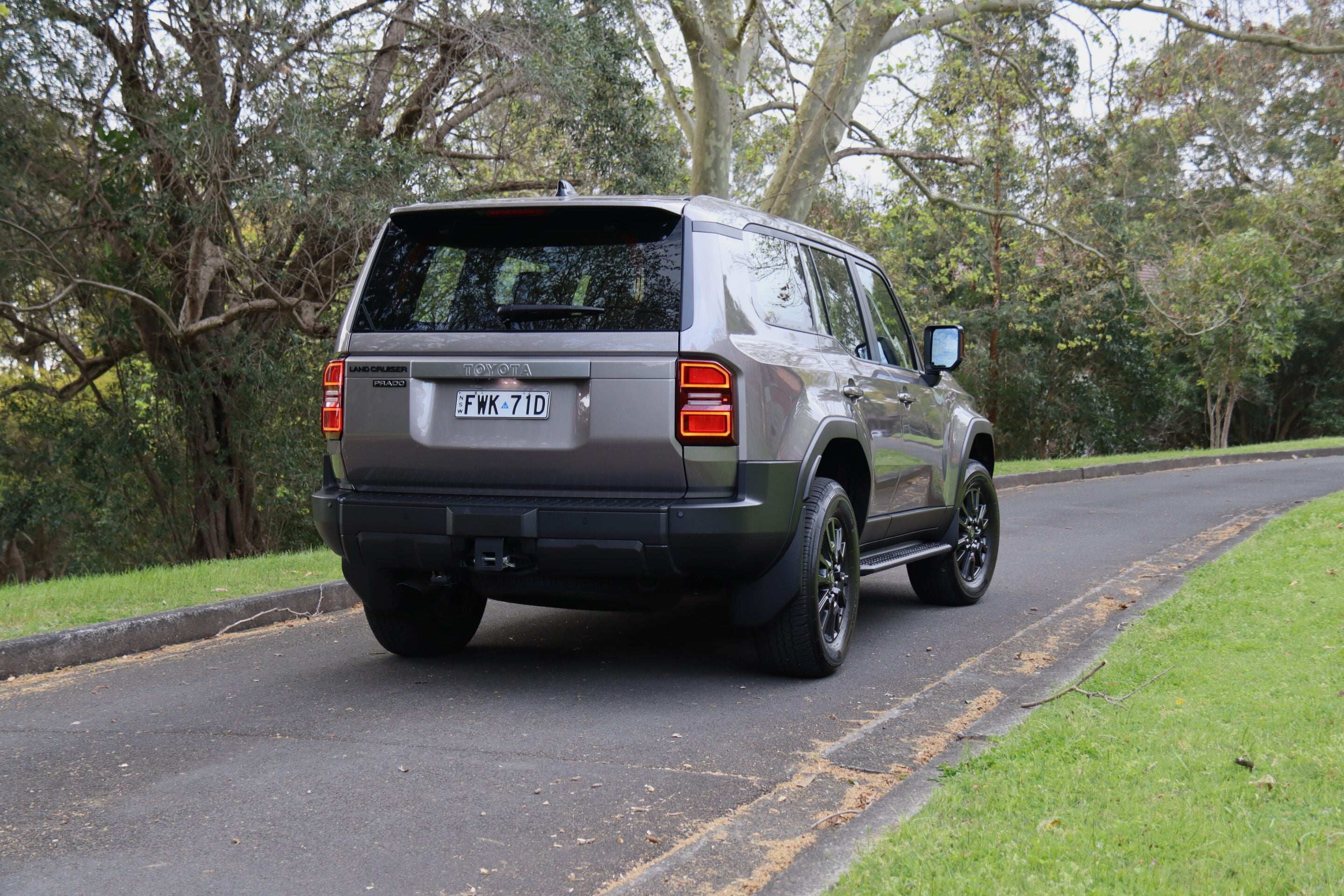 Rear three-quarter view of a gray Toyota Land Cruiser Prado parked on a residential street with trees in the background.