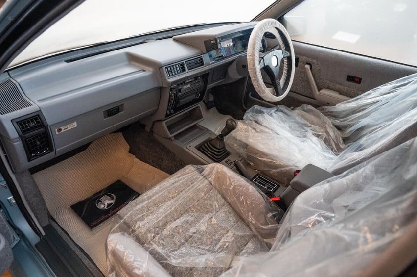 Left-hand drive car interior with seats wrapped in plastic, gray dashboard, and a manual gear shift.