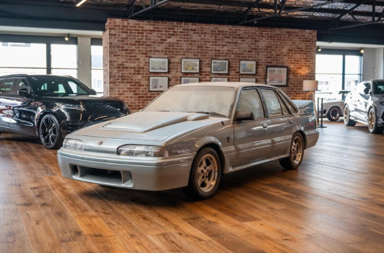 Silver vintage sedan with a pronounced hood scoop showcased on a wooden showroom floor, brick wall with framed pictures in the background.