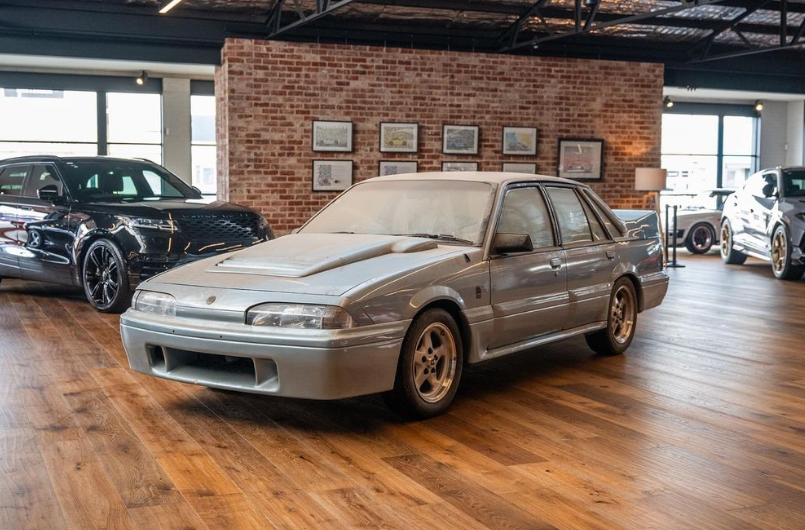 Silver vintage sedan with a pronounced hood scoop showcased on a wooden showroom floor, brick wall with framed pictures in the background.